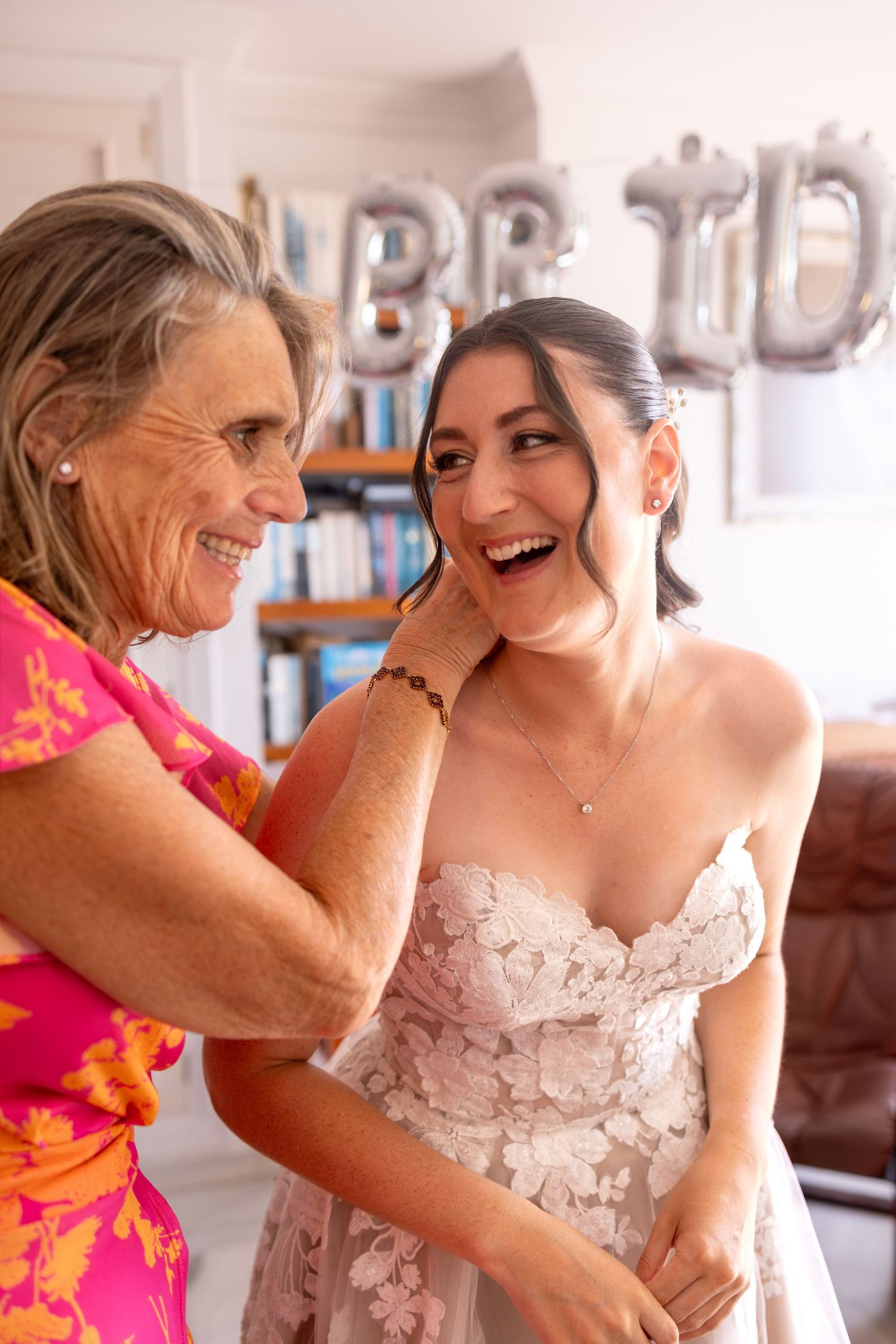 A joyful bride in a floral gown laughs with an older woman in a pink dress during wedding preparations.