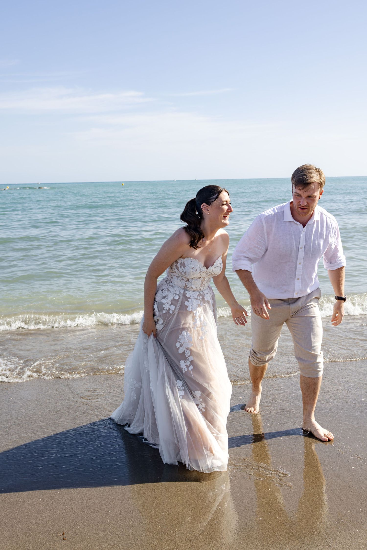 Joyful bride and groom laugh together while walking barefoot along the shoreline on their wedding day.