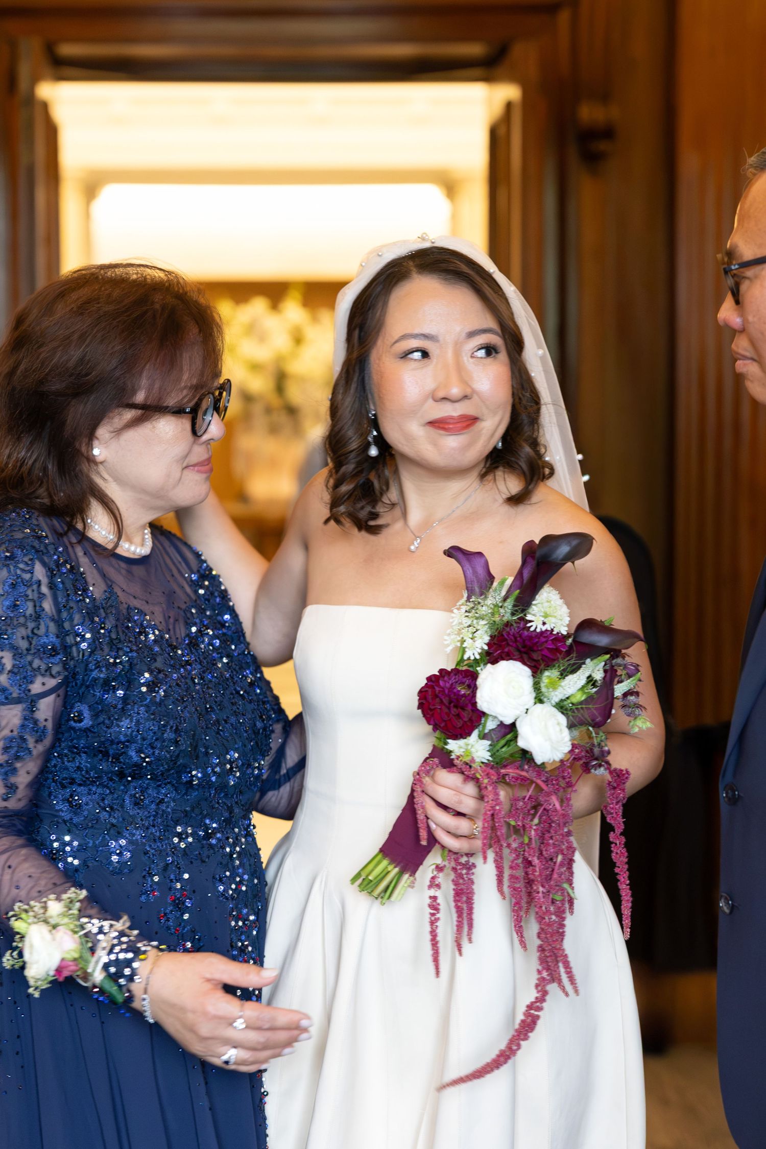 Elegant bride in strapless white gown holds bouquet of red roses and calla lilies flanked by family members.