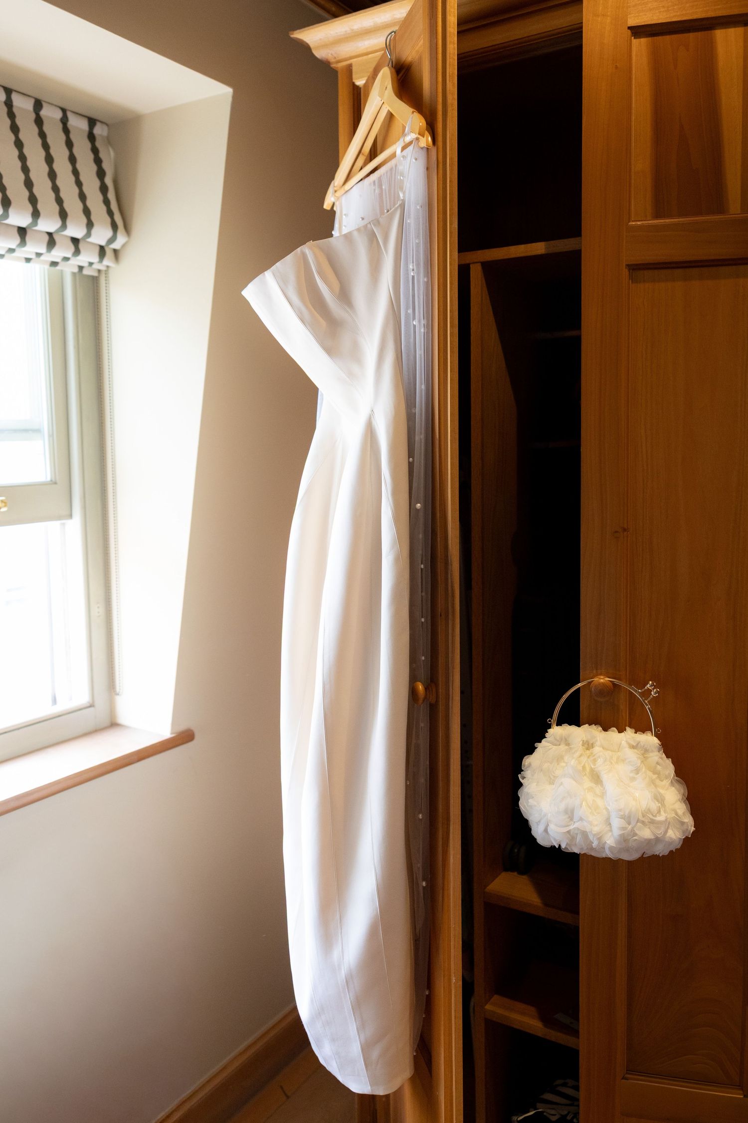 White wedding dress hanging on wooden wardrobe with decorative bridal purse, beside a window in a bright room.