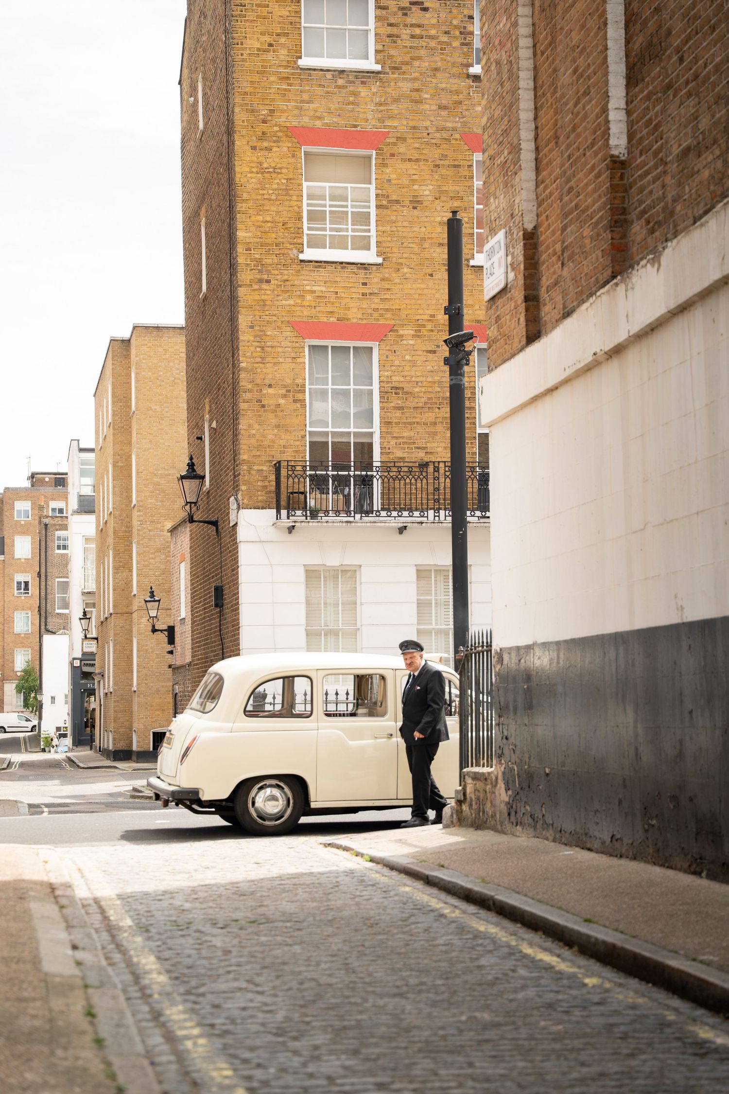 A chauffeur in uniform stands beside a vintage cream-colored car on a cobblestone London street.