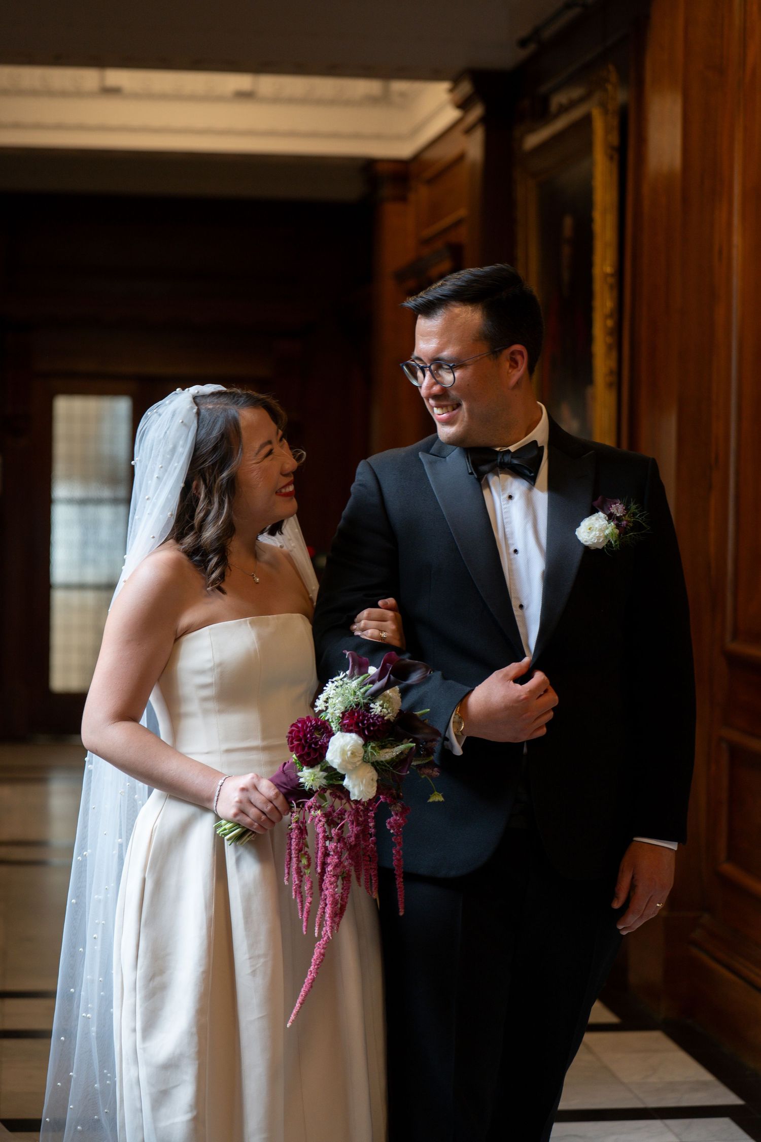 Bride and groom share a joyful moment, she in a strapless gown with veil, he in a tuxedo, holding a burgundy bouquet.