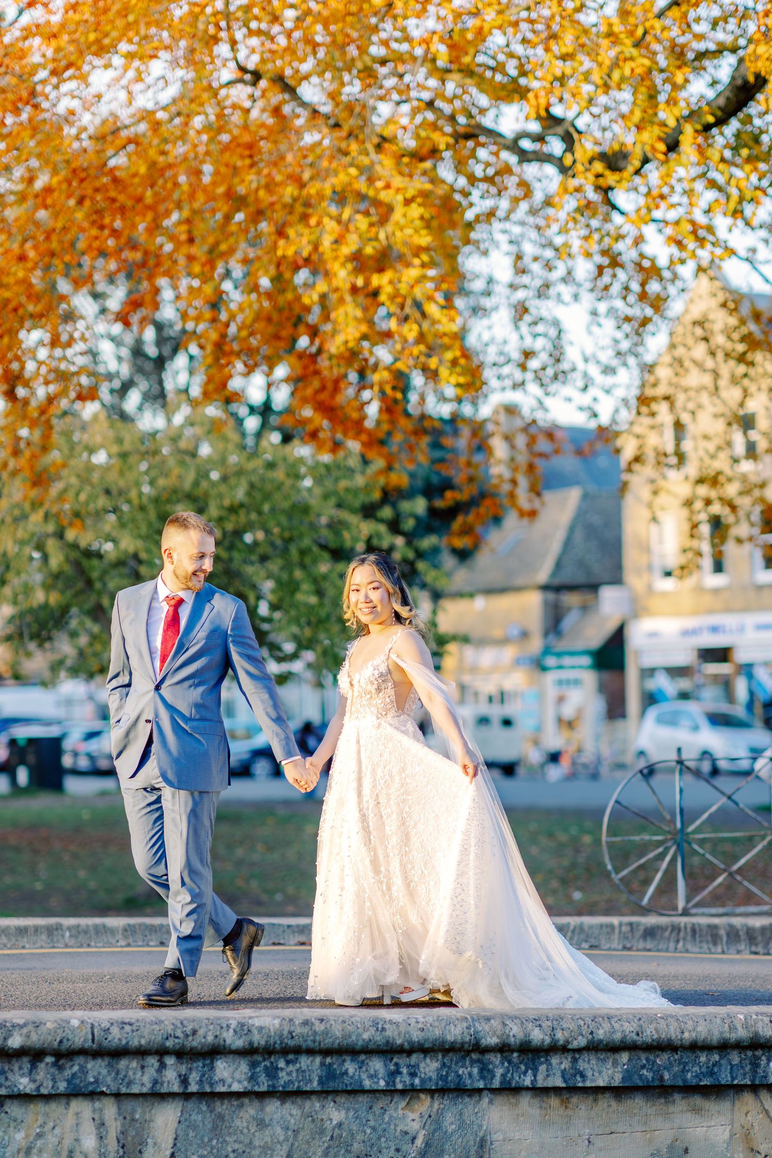Wedding couple walking along stone wall under vibrant autumn foliage.
