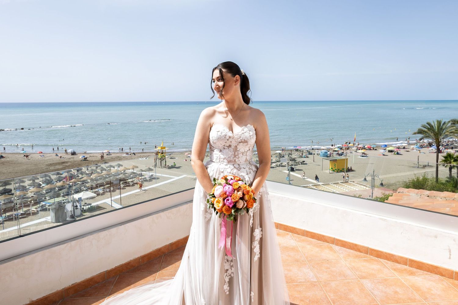 Bride in floral gown holds colorful bouquet on rooftop terrace overlooking a sunny beach and blue sea.