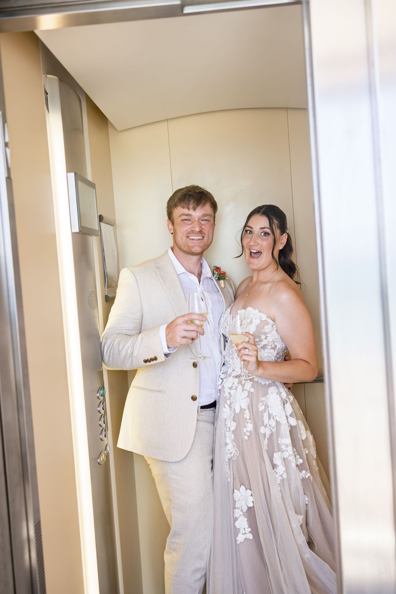 Bride and groom holding champagne flutes in an elevator, smiling joyfully on their wedding day.