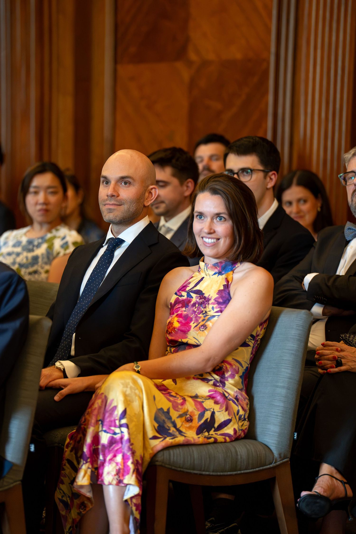 Elegantly dressed wedding guests seated in wooden paneled venue, woman in floral dress smiling.