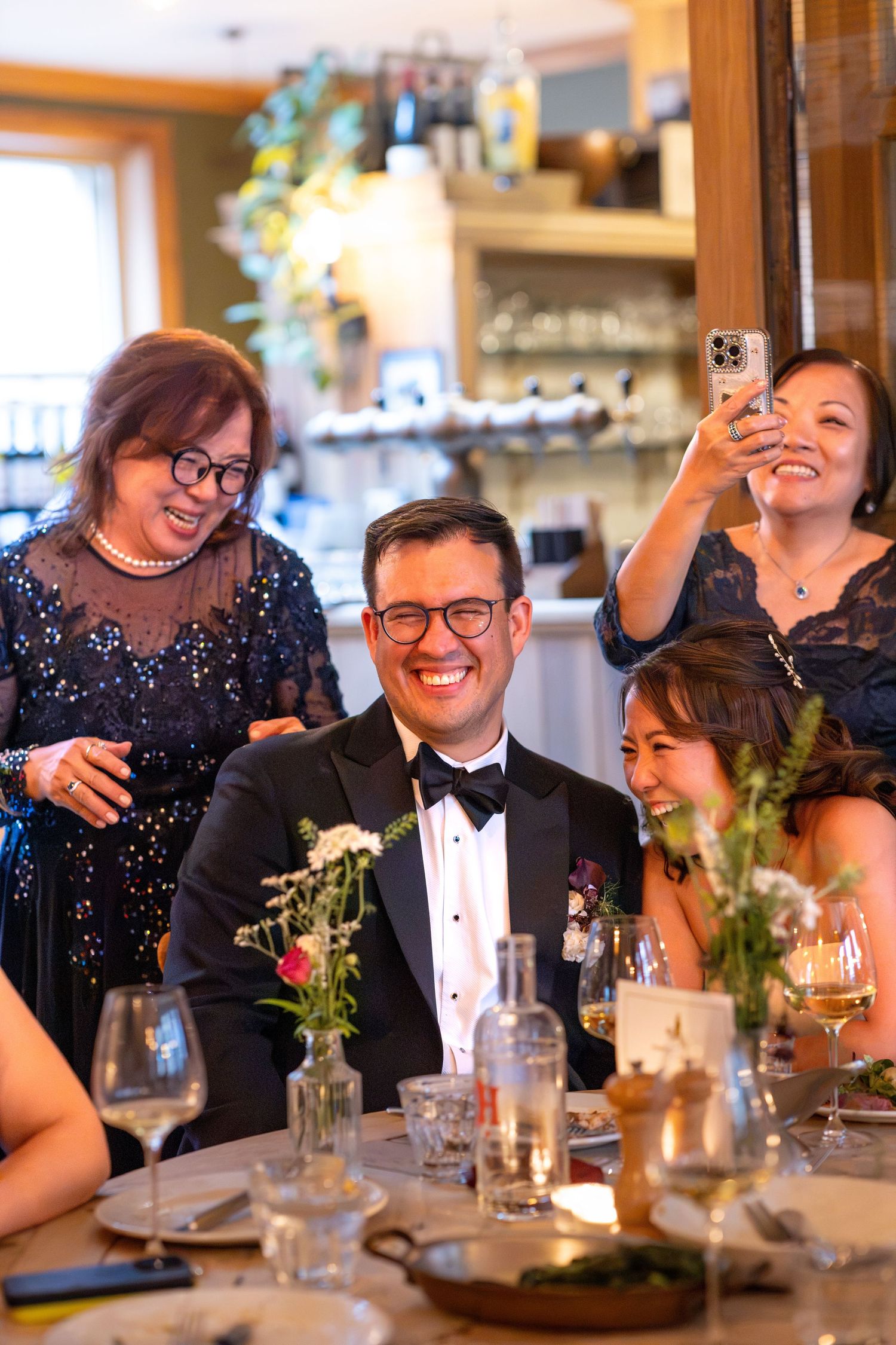 Groom in tuxedo laughs with family and guests at elegant wedding reception dinner table.