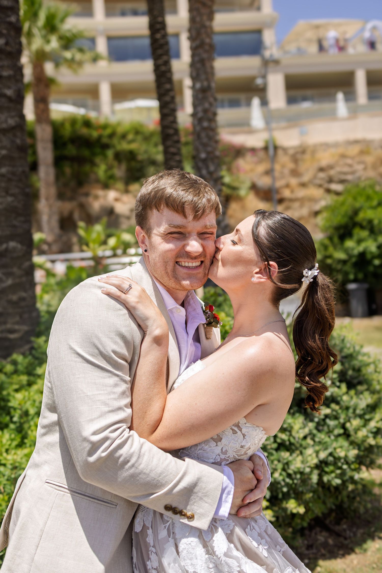 Smiling groom being kissed on the cheek by bride in a lush garden setting at a resort wedding.