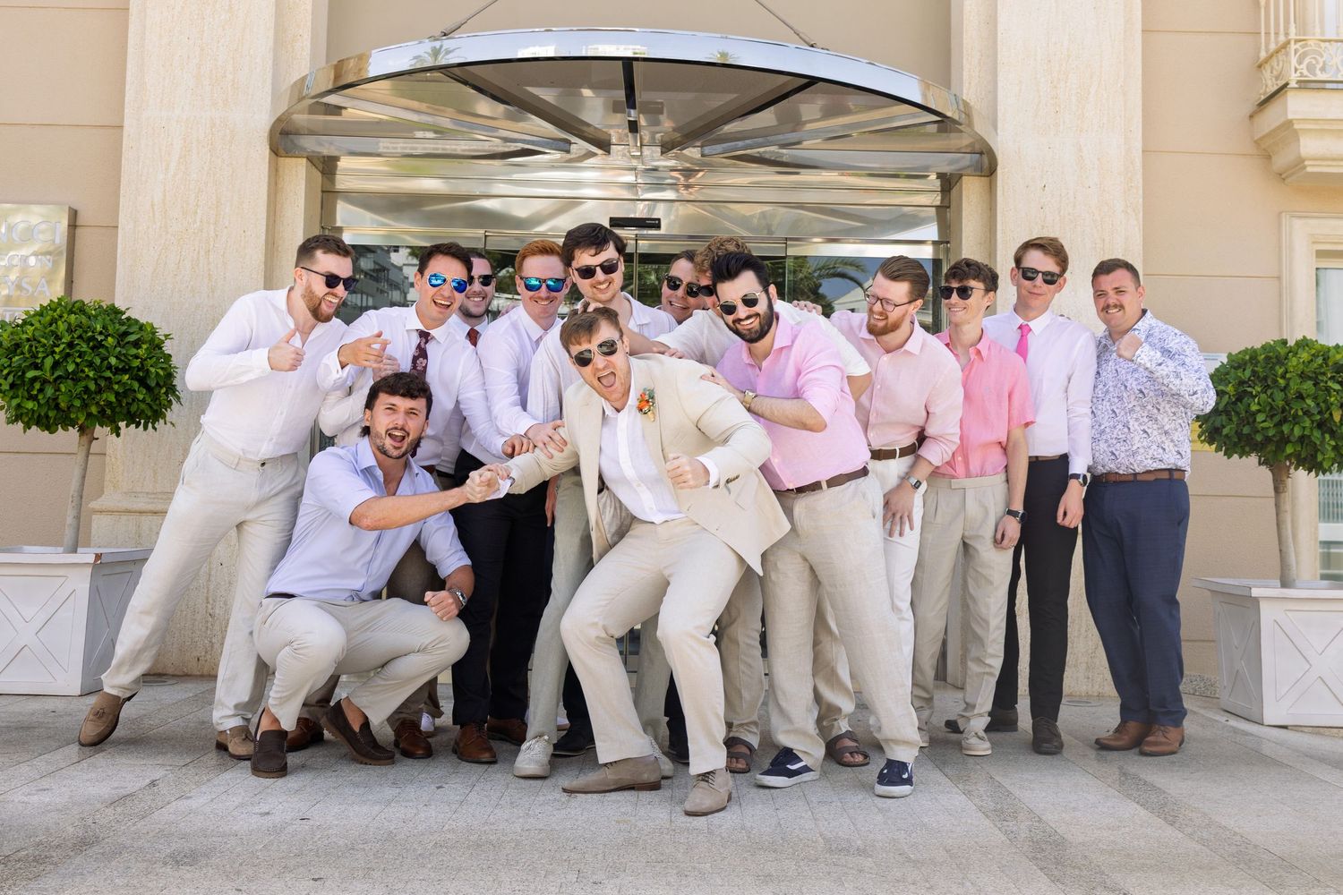 Groomsmen in pink shirts and khakis posing playfully outside a building on a sunny wedding day.