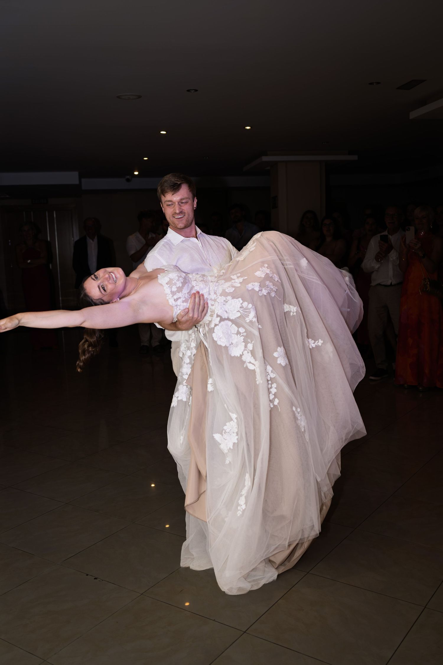 Groom lifts bride in floral lace gown during energetic first dance at wedding reception.