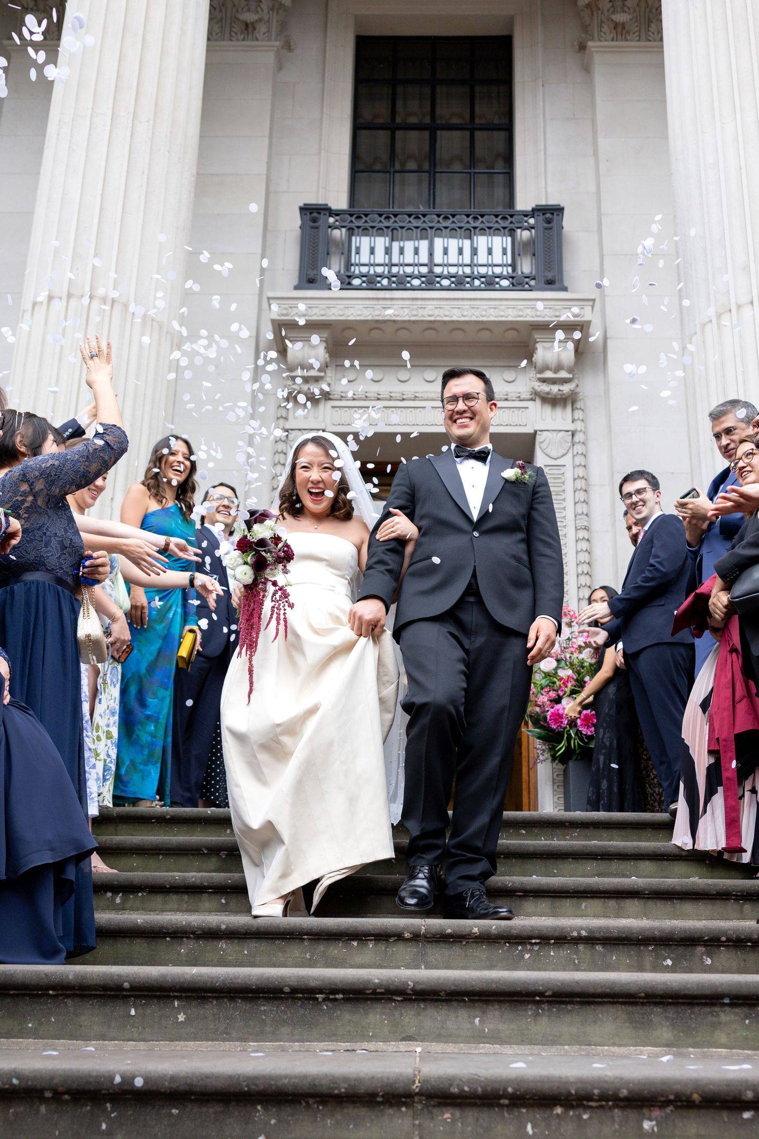 Smiling newlyweds walk down grand stone steps through shower of white confetti thrown by guests.
