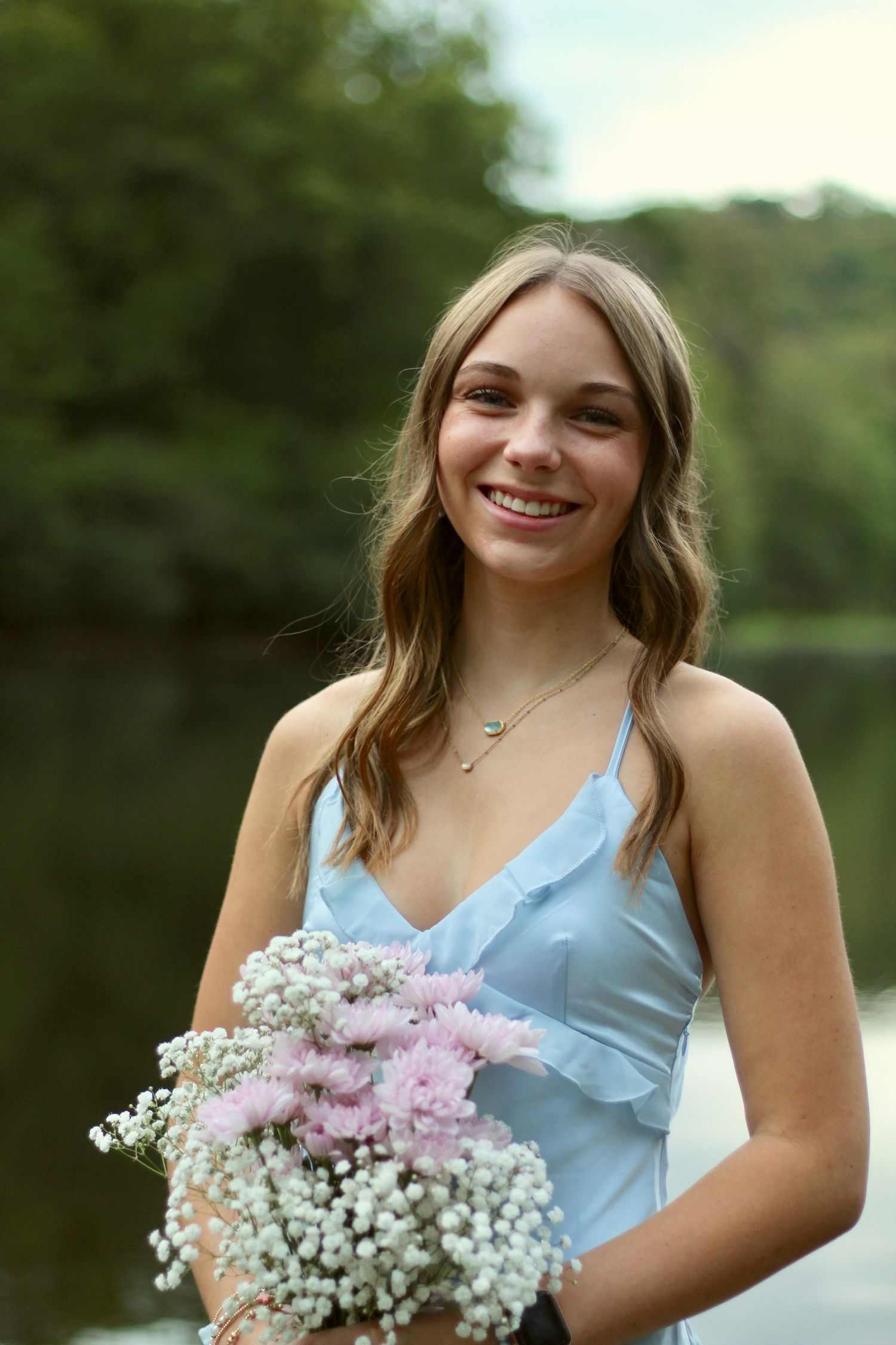 In her blue gown, in front of a pond, she smiles at the camera holding her bouquet.