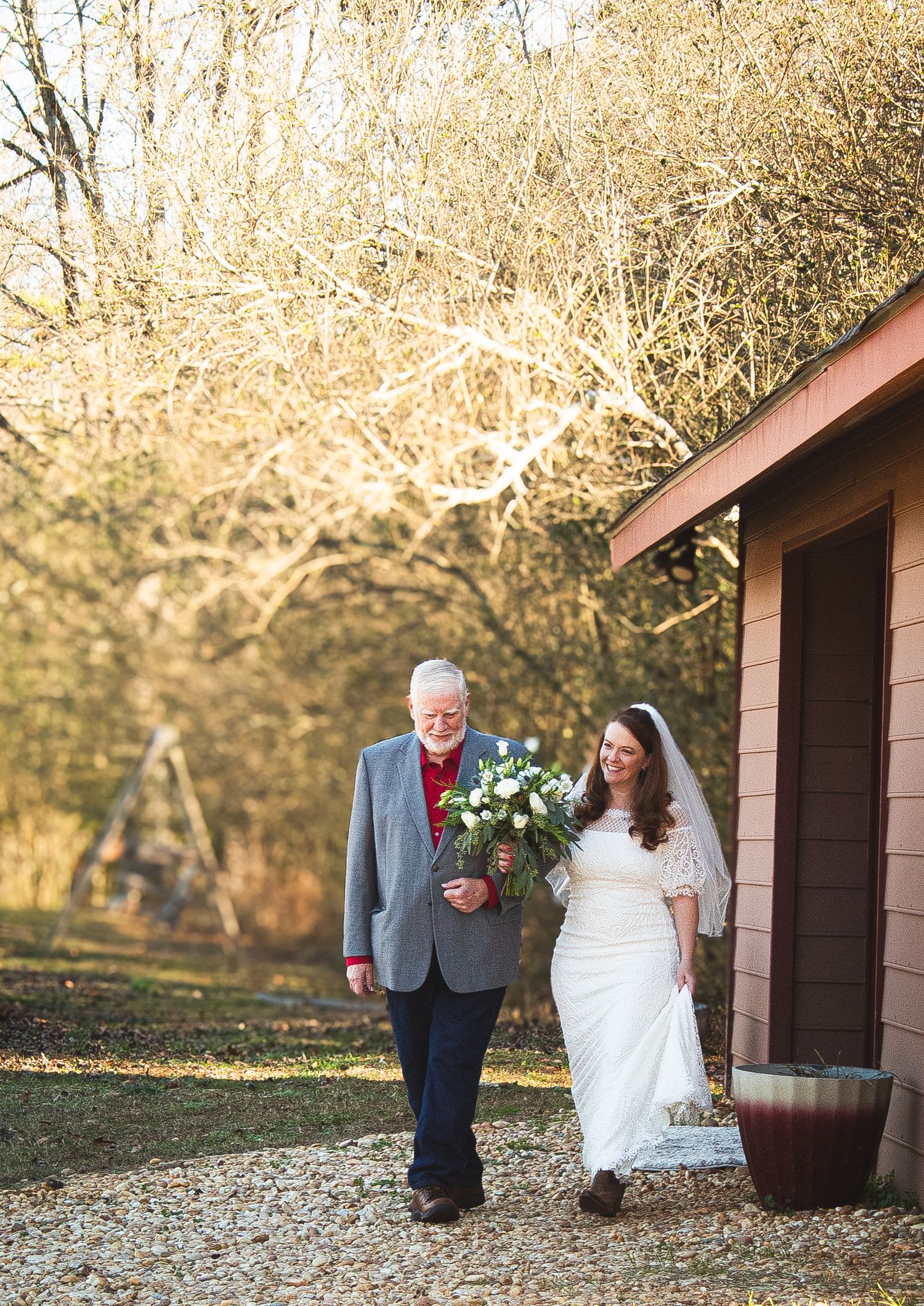 Bride and her dad walking toward the aisle with trees behind her.