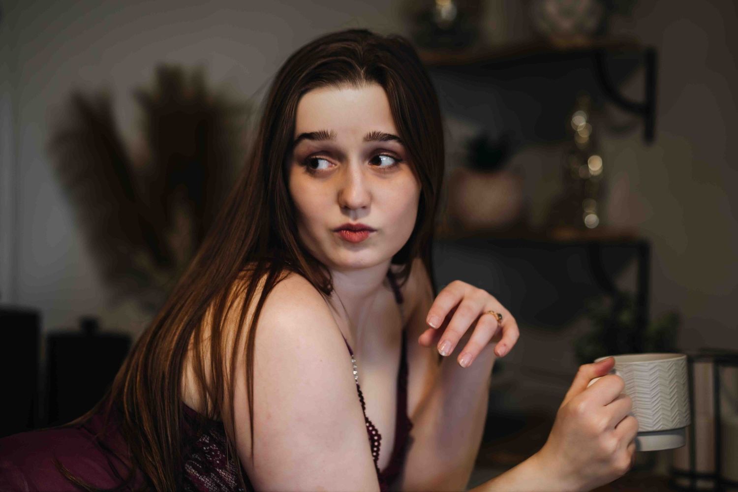 Young woman with long brown hair holding a white mug, looking thoughtful in a cozy dimly lit room.