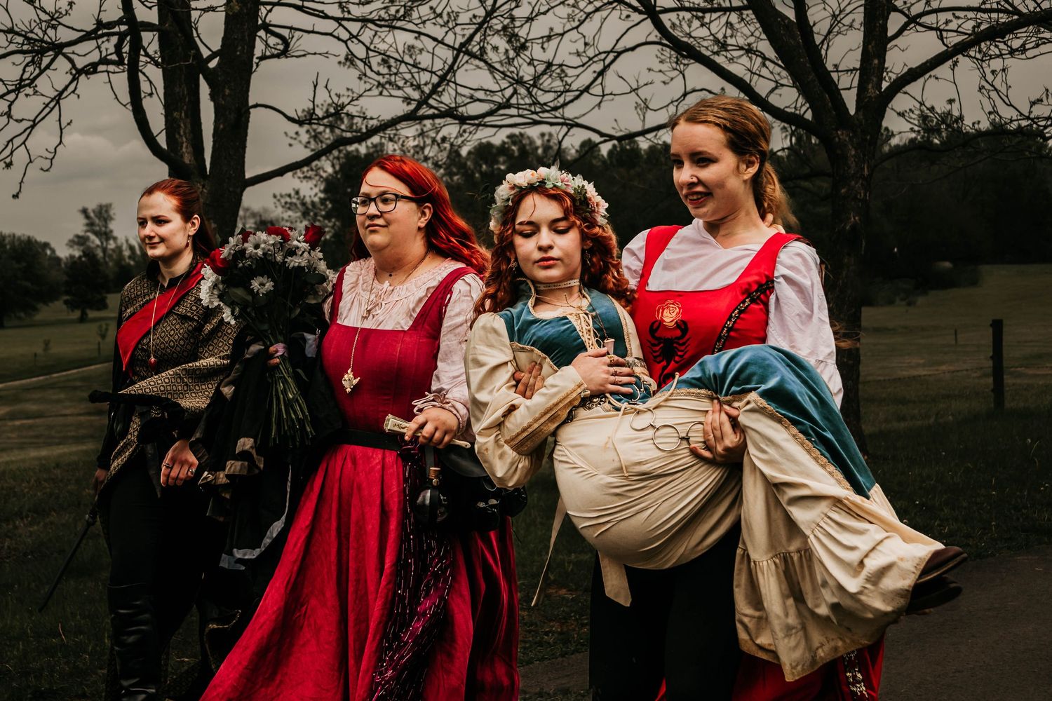 Wedding party wearing red and beige dresses poses together in a dramatic outdoor setting at dusk.