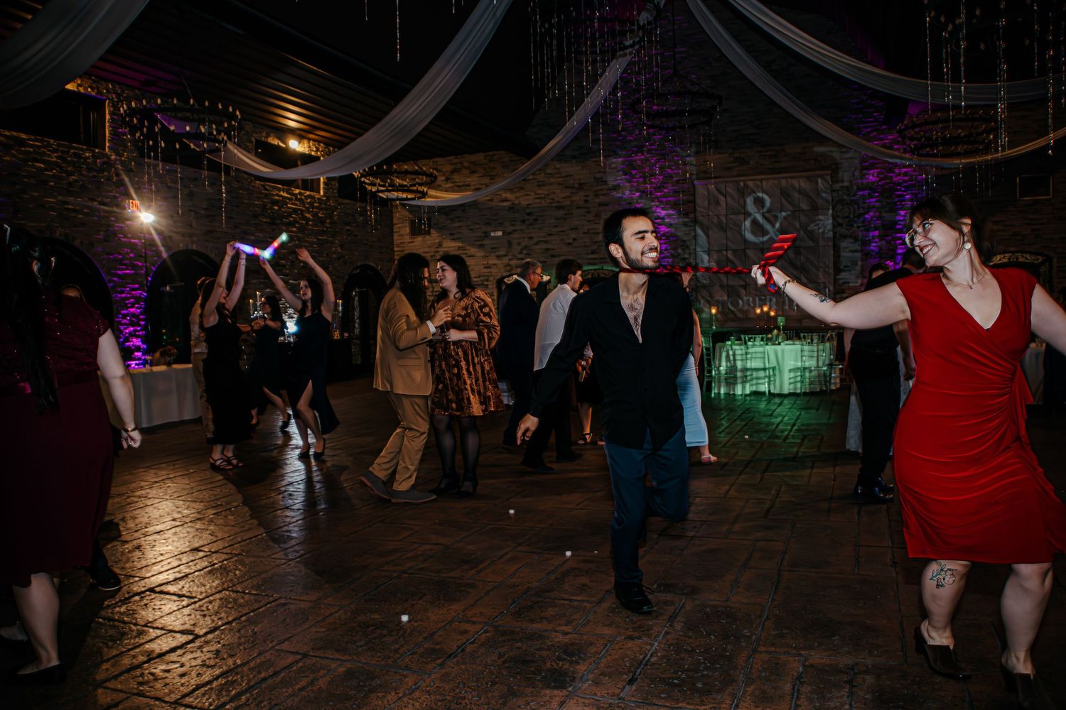 People dancing and celebrating at a dimly lit wedding reception with purple and green lighting effects on the dance floor.