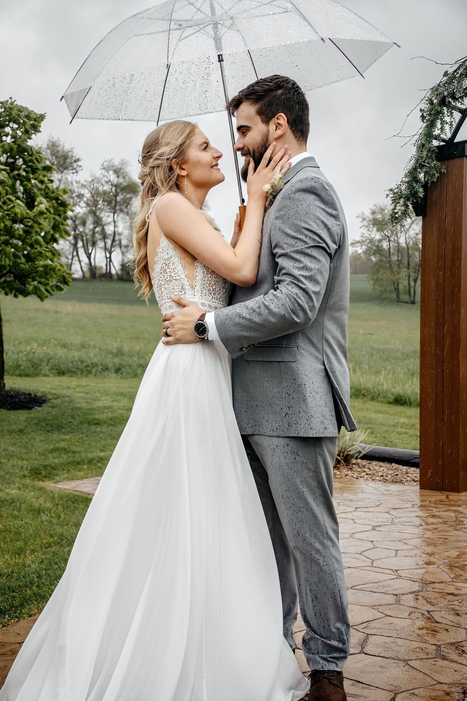 A bride and groom share an intimate moment under a clear umbrella on a rainy wedding day in a garden setting.