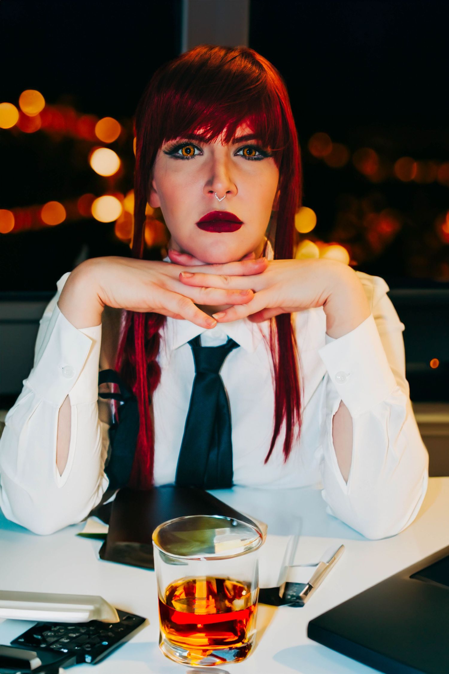Portrait at night with red hair, white blouse, black tie, and whiskey glass on table with laptop in dark setting.