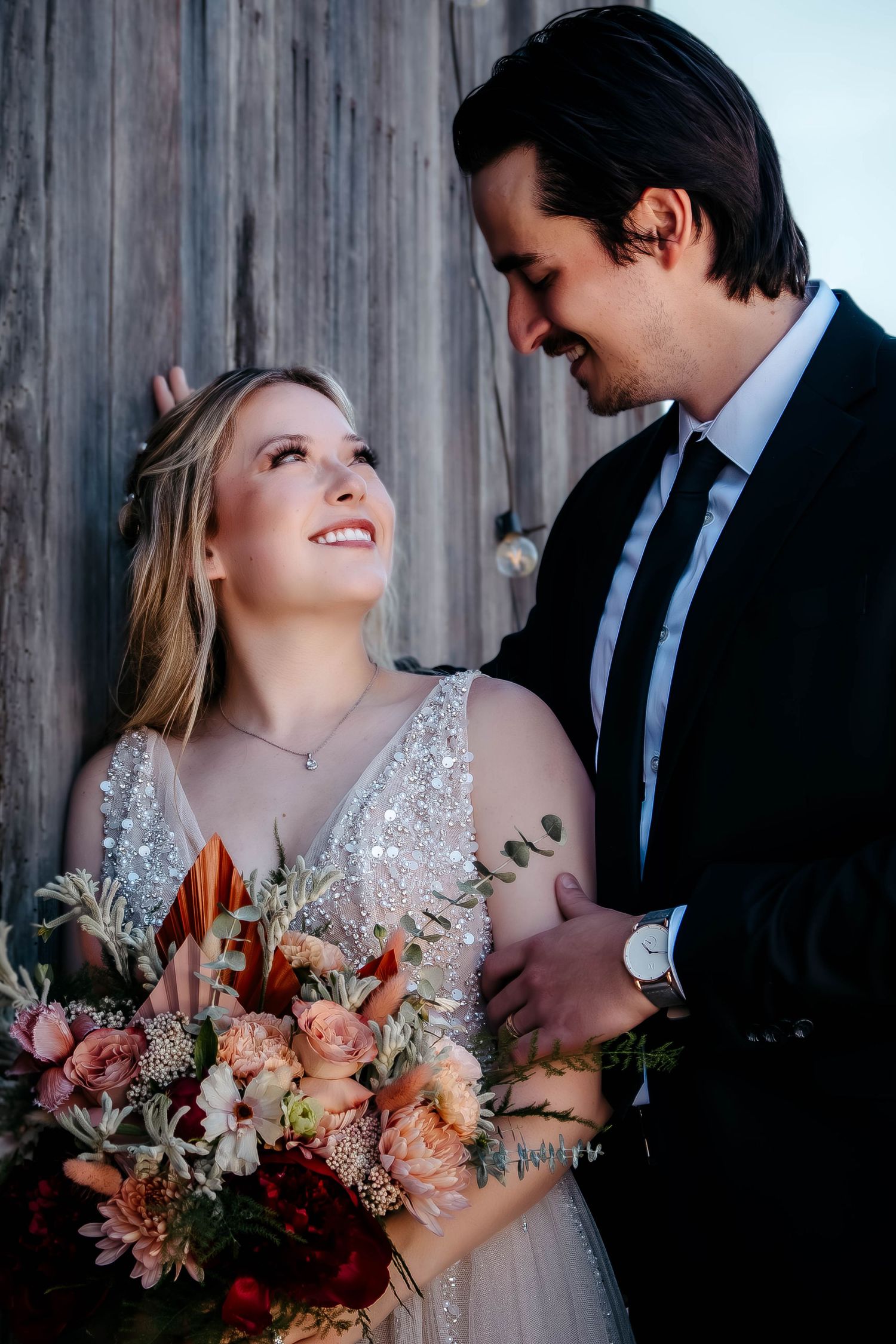 Bride in lace dress holding colorful bouquet gazes lovingly at groom against rustic wooden wall backdrop.