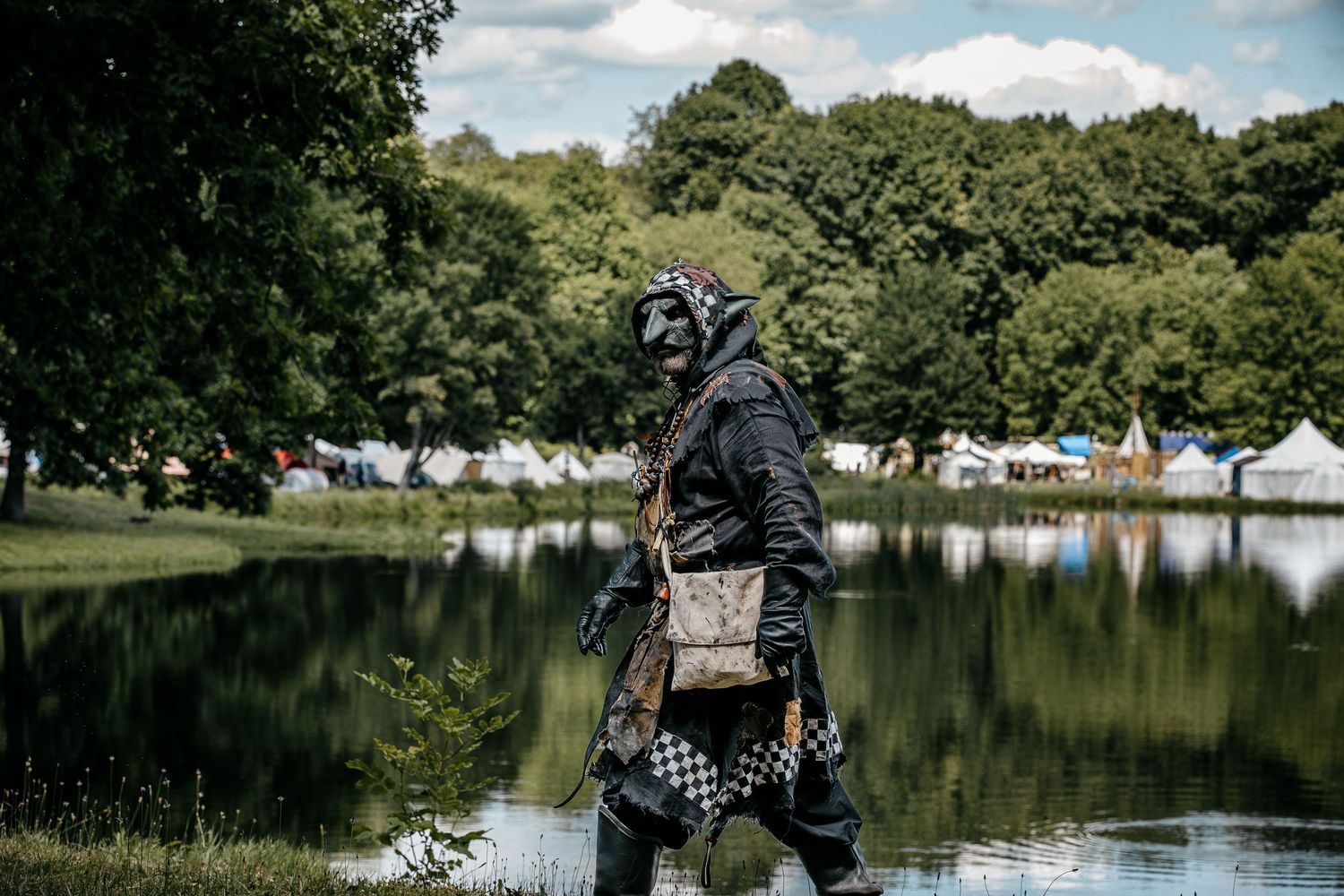 Person in dark post-apocalyptic costume and gas mask walking beside a lake at an outdoor festival.