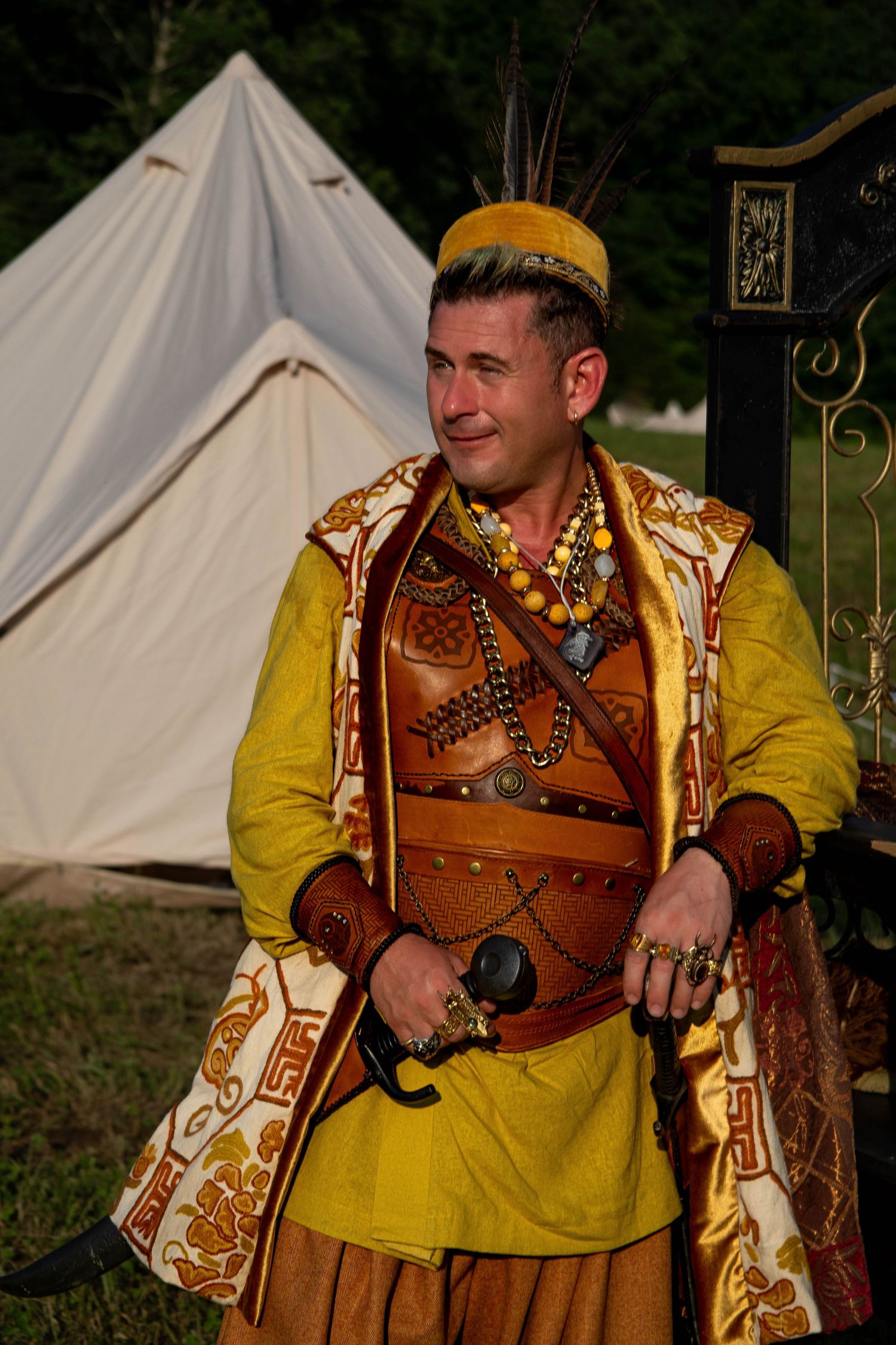 Medieval performer in yellow tunic and brown leather armor with ornate decorations poses near white canvas tents.