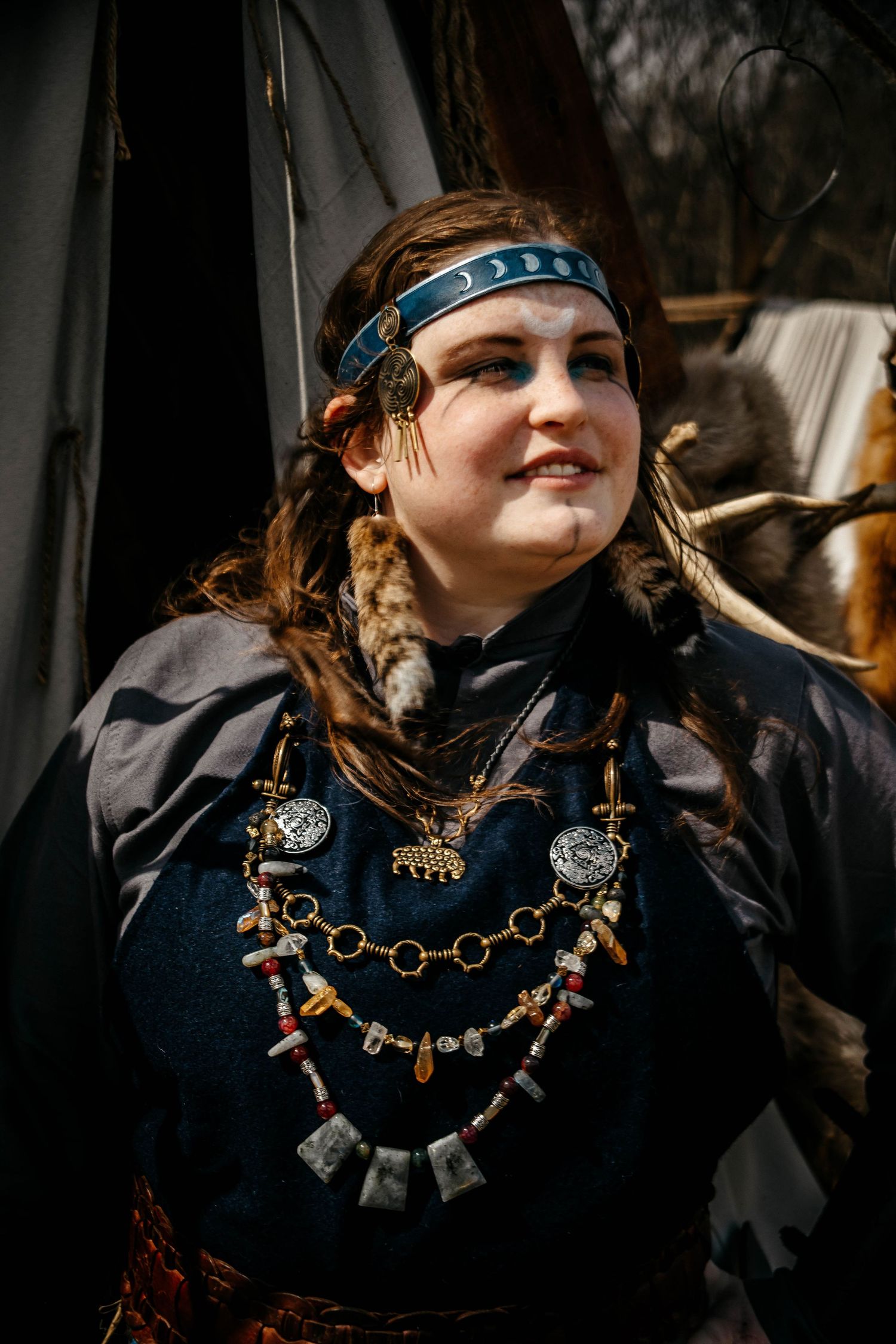 Medieval style portrait showing decorative jewelry with stones and charms worn over dark clothing with a blue headband.