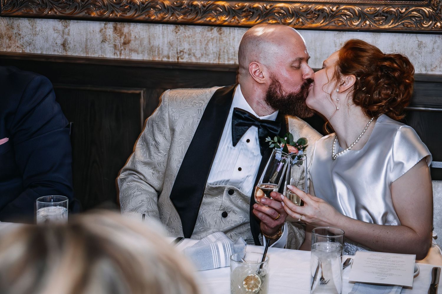 Bride and groom sharing a romantic kiss at wedding reception table, holding champagne flutes in elegant venue.