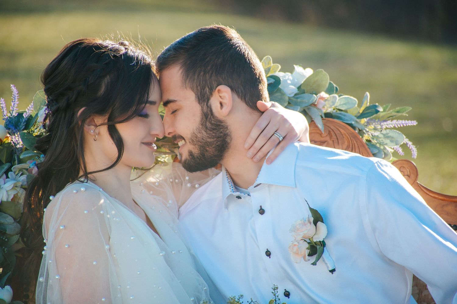 A bride and groom share a romantic almost-kiss at their outdoor wedding, surrounded by a floral arrangement.