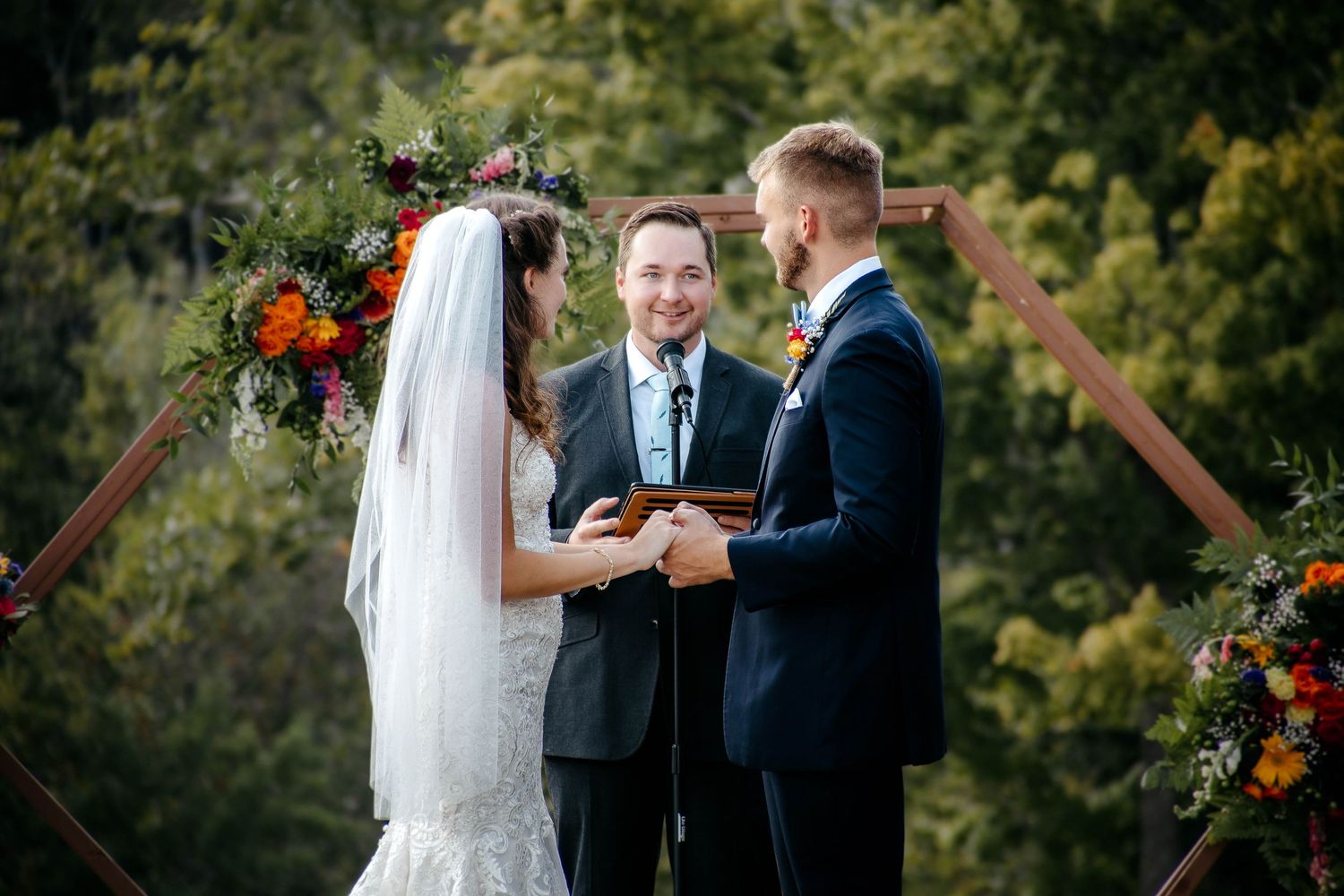 Bride and groom exchange vows at outdoor wedding ceremony beneath floral arch with officiant and lush greenery backdrop.