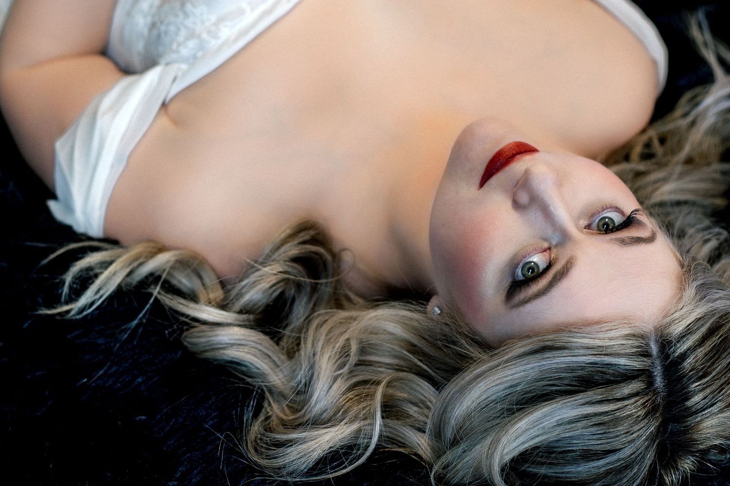 A young woman with blonde wavy hair and red lips lying down, gazing upward in a dramatic portrait pose.