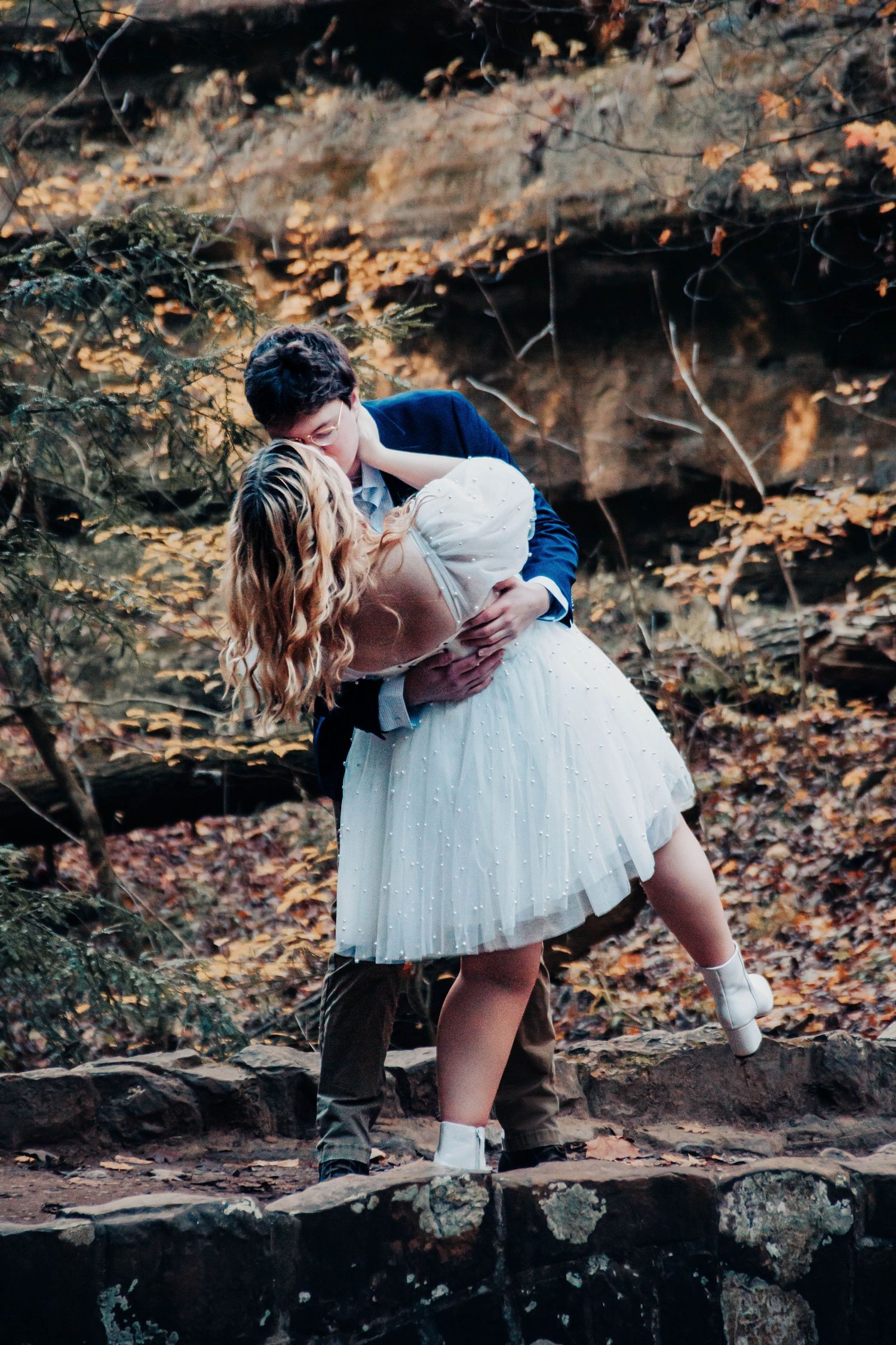 A couple in romantic attire shares an intimate moment on rocky terrain surrounded by fall leaves.