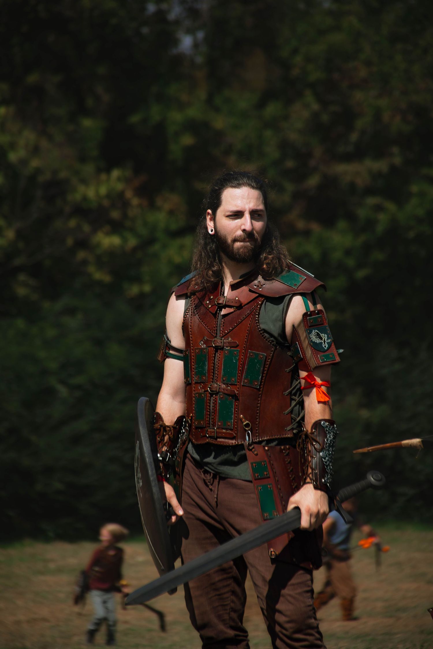 A person in medieval leather armor and costume holds a sword while participating in an outdoor combat reenactment event.