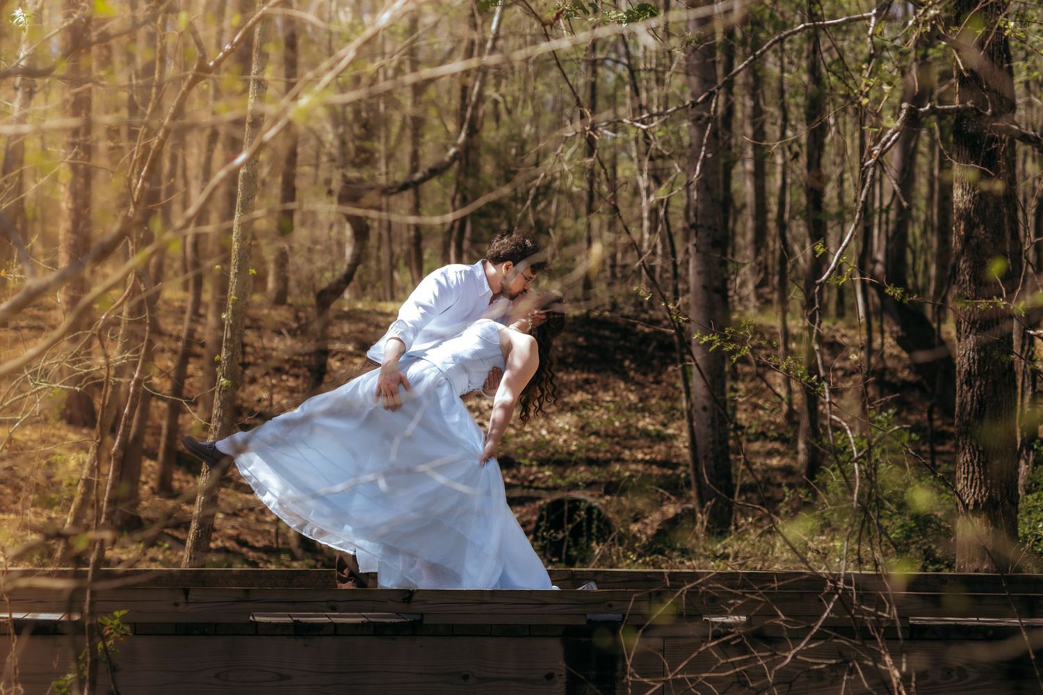 A bride and groom share a romantic dip kiss on a wooden bridge surrounded by a sunlit forest.