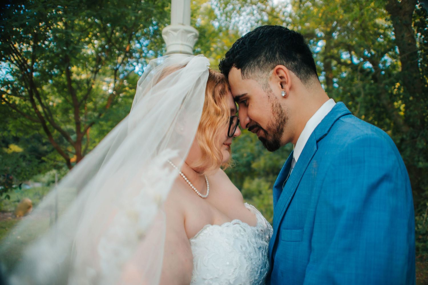 A couple shares an intimate moment in their wedding attire, with the bride in white and groom in blue against a natural backdrop.
