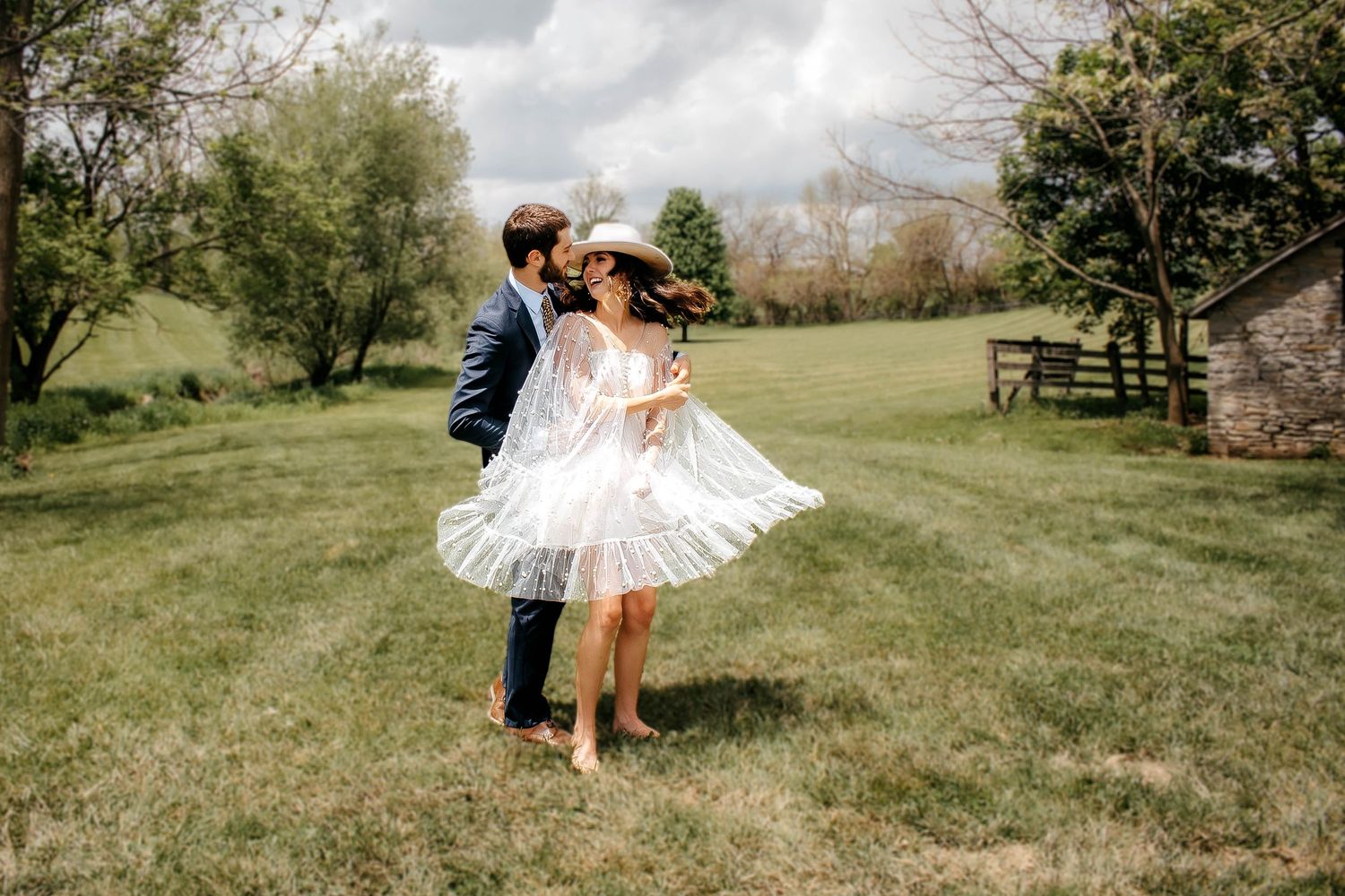 A couple shares an intimate moment together in a scenic green field with rustic buildings in the background.