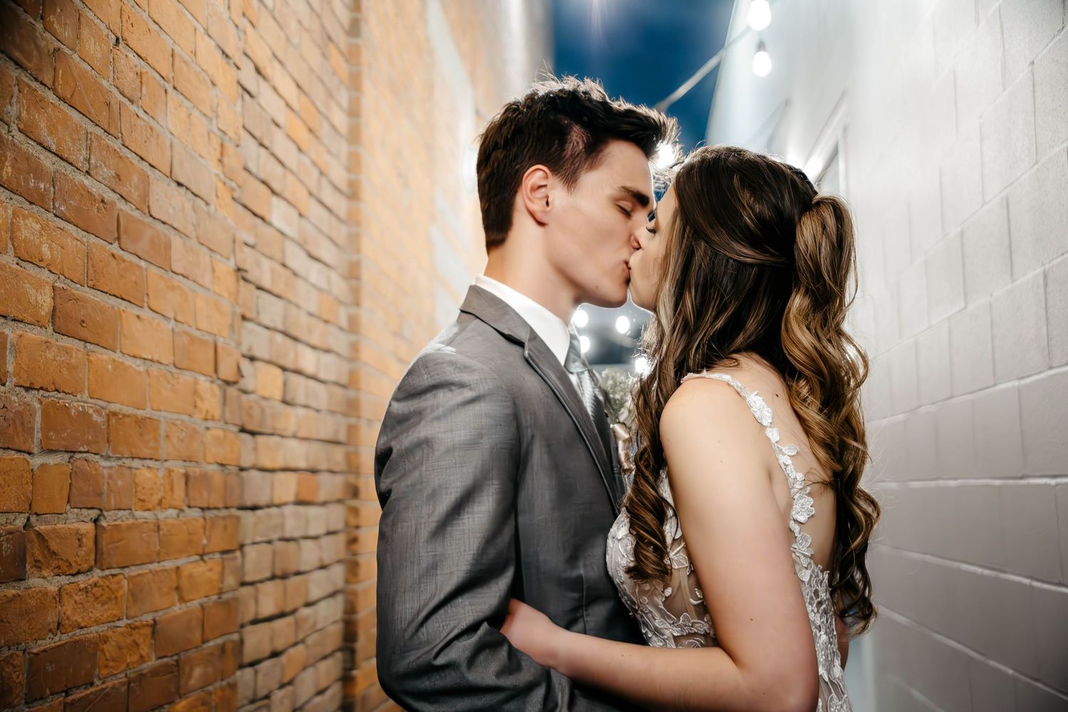 Romantic couple in formal attire sharing intimate moments against brick wall in wedding photo series.
