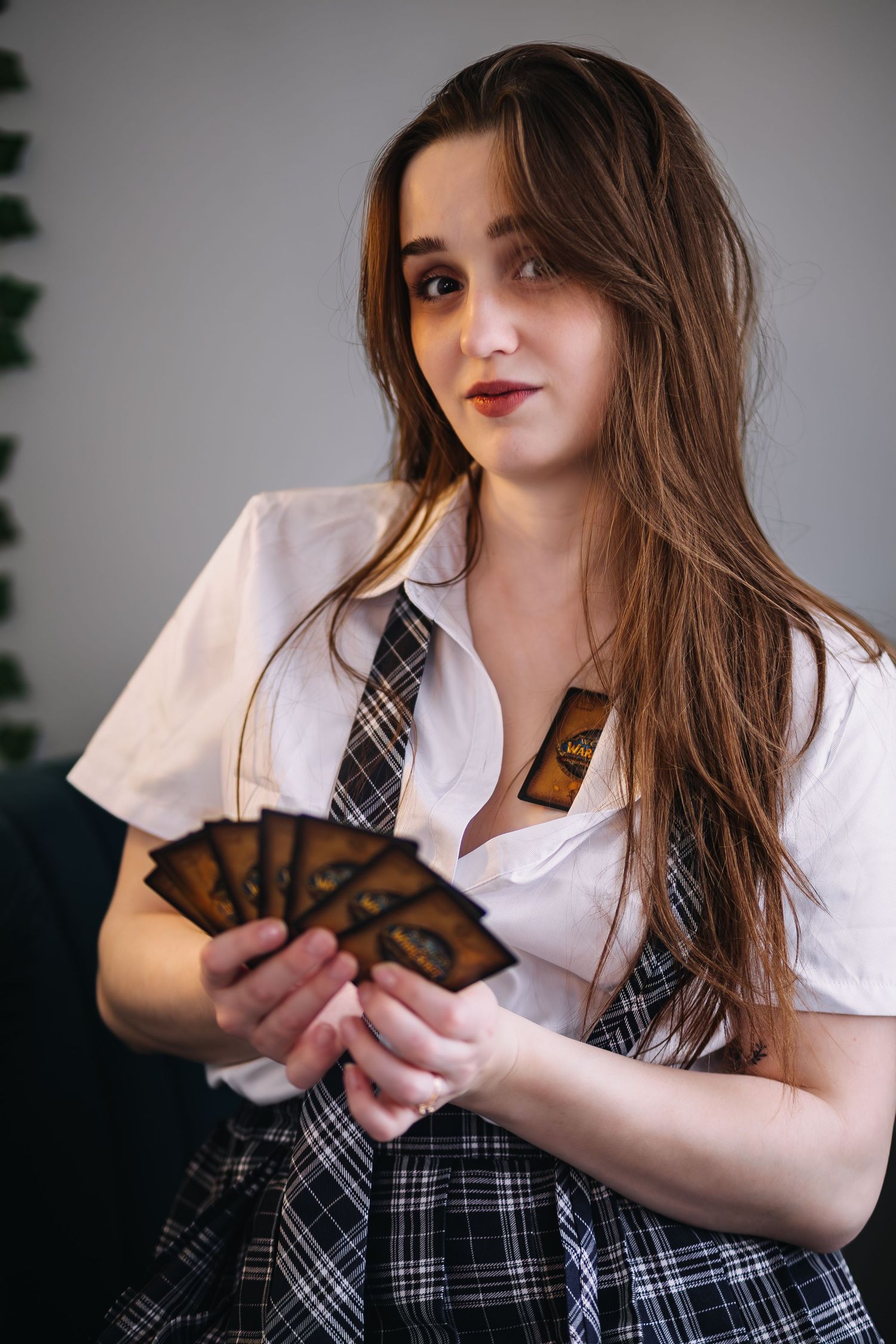Young woman in school uniform holding fan of trading cards, posing indoors against gray background.