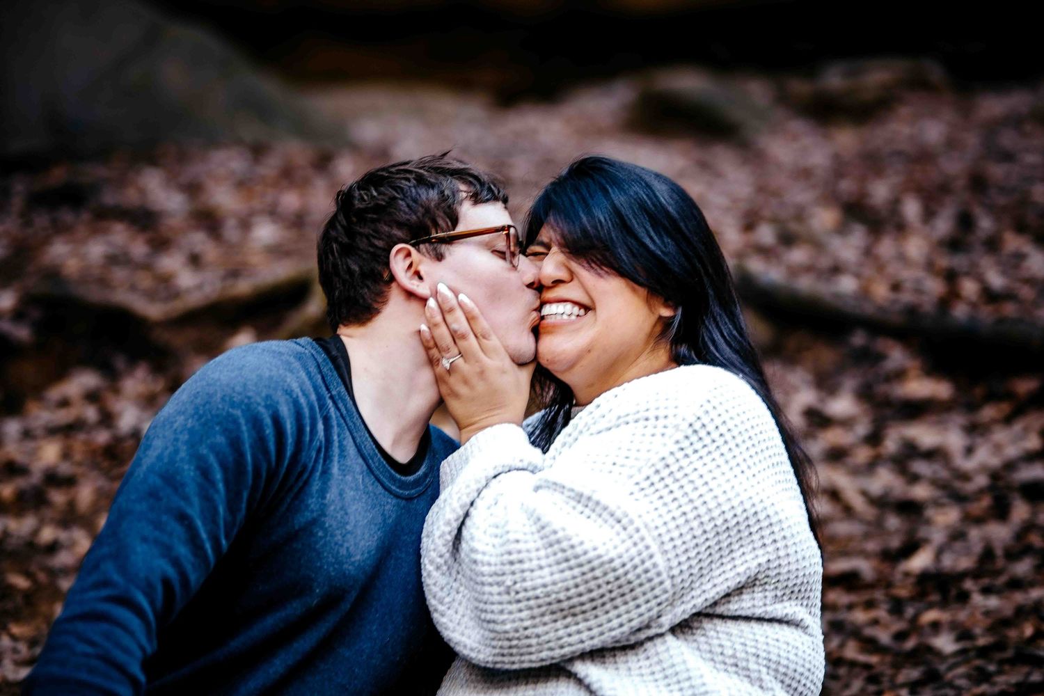 Couple sharing a romantic kiss outdoors in autumn woods, man gently holding woman's face as she smiles.