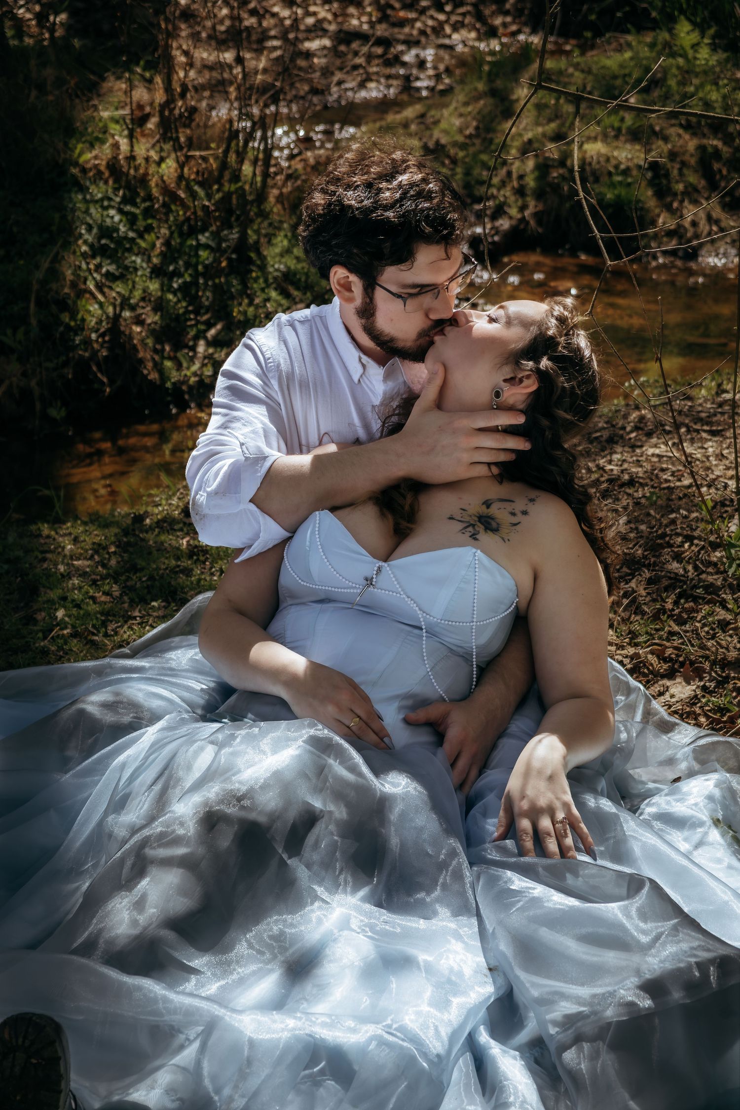 A couple shares a romantic kiss outdoors, with the woman in a flowing blue gown reclining against her partner.