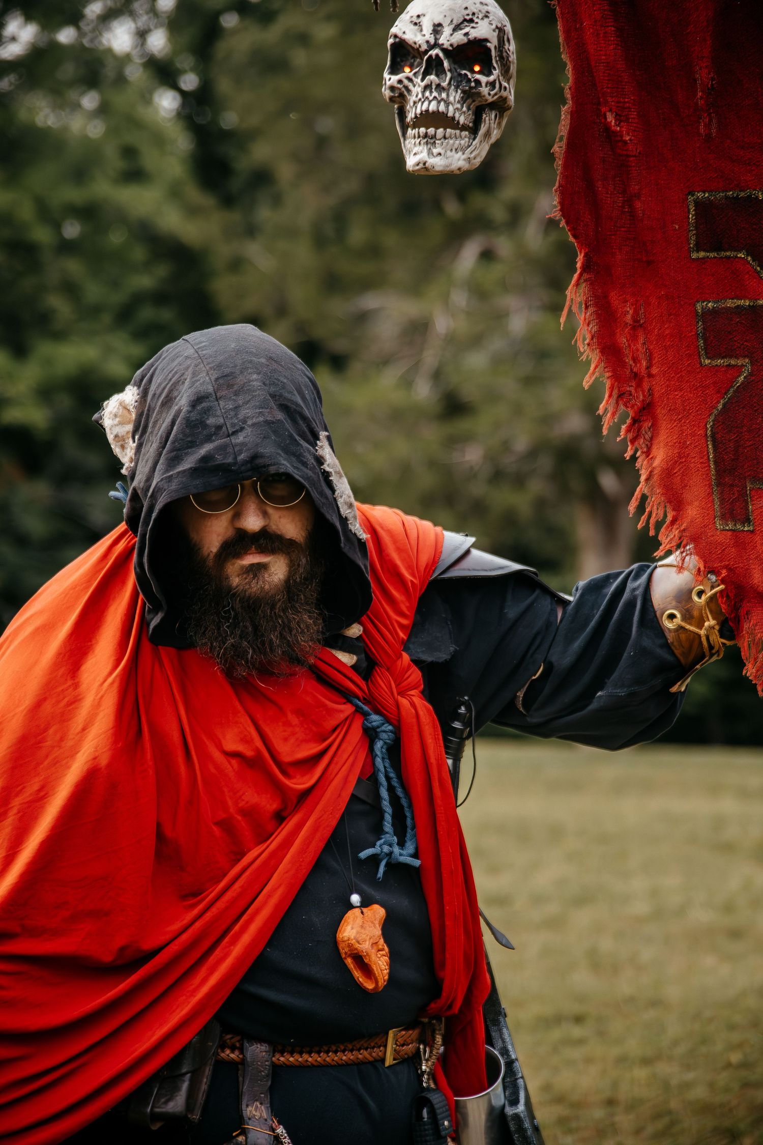 Bearded man in medieval fantasy costume with red cape, dark hood, and skull prop at outdoor LARP event.