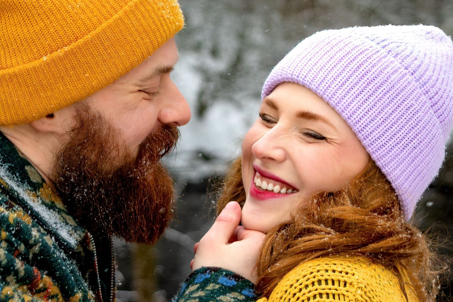 A couple wearing winter hats and scarves laughs together during a snowy outdoor photo session.
