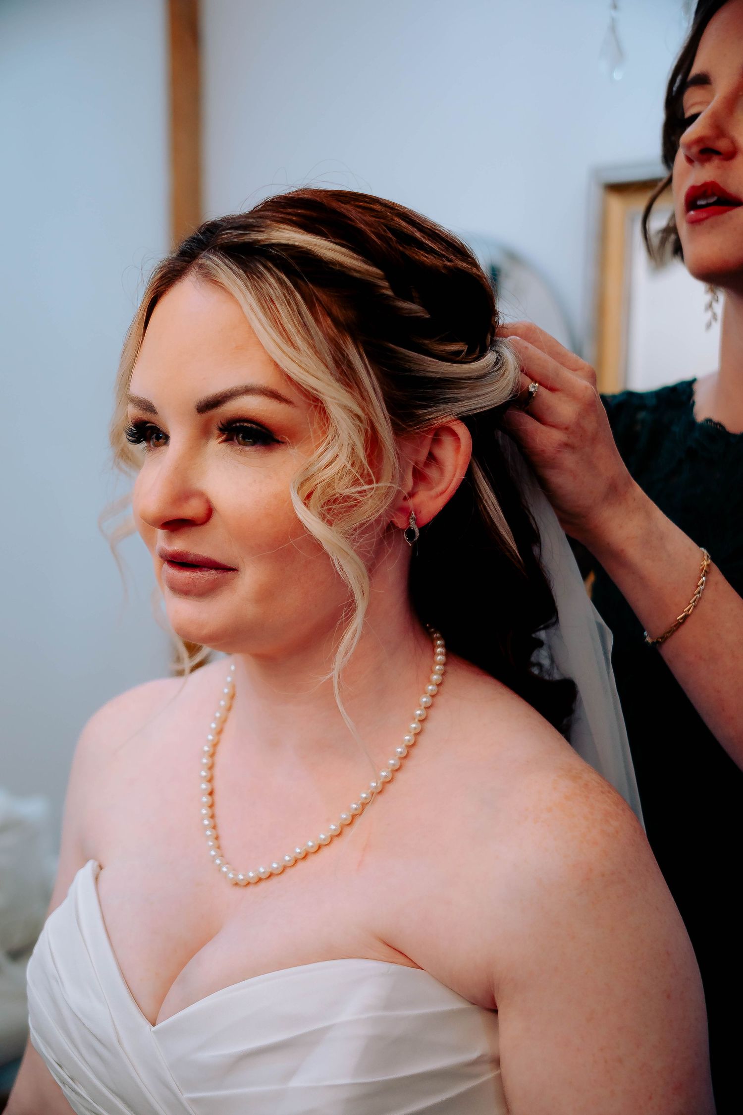 A bride wearing a white strapless dress and pearl necklace gets her hair styled on her wedding day.