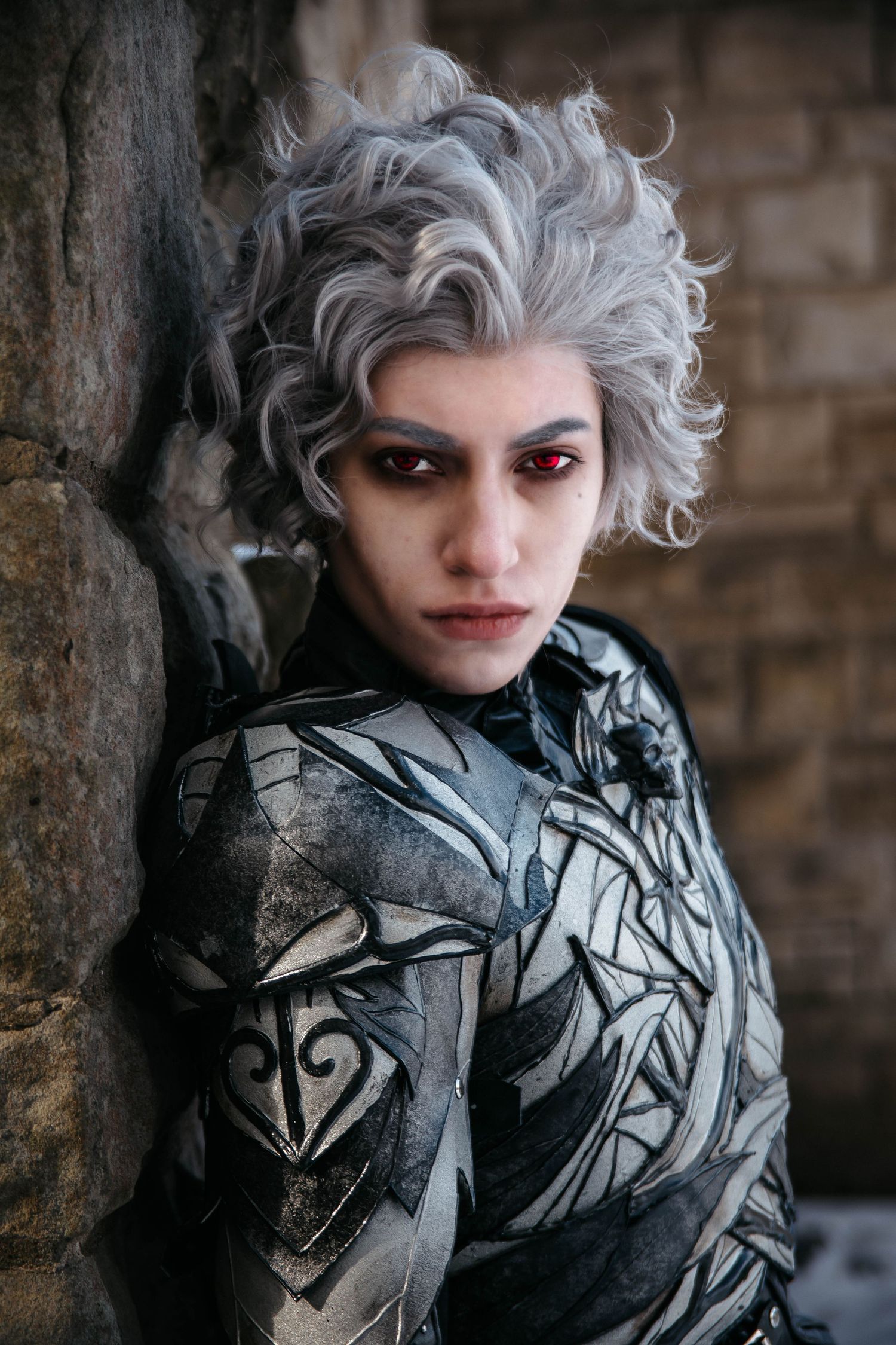 Dramatic portrait of someone in silver-gray costume makeup and dark clothing against a brick wall.