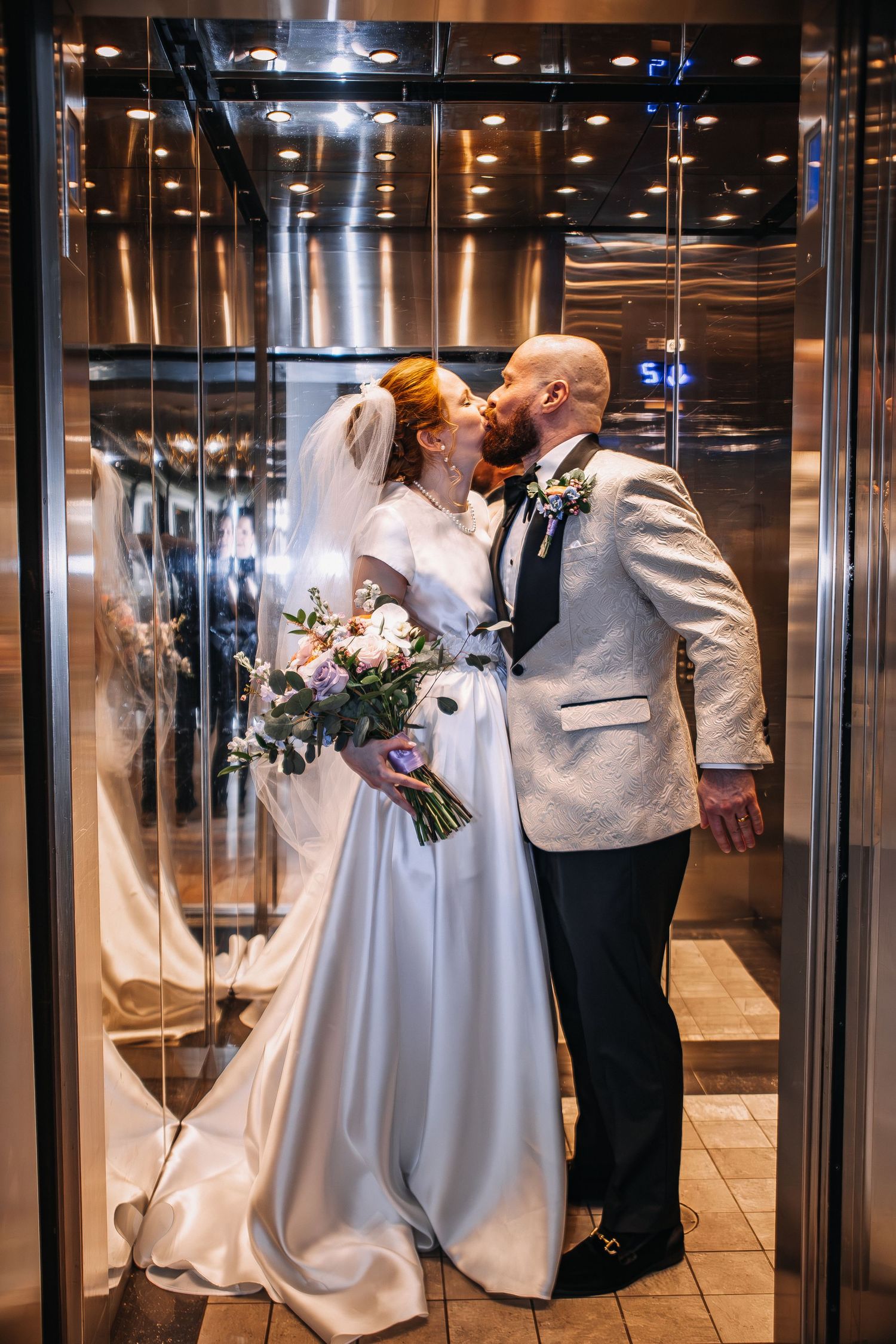 Bride and groom sharing a romantic kiss in a elegant mirrored elevator, bride holding a colorful bouquet in wedding attire.