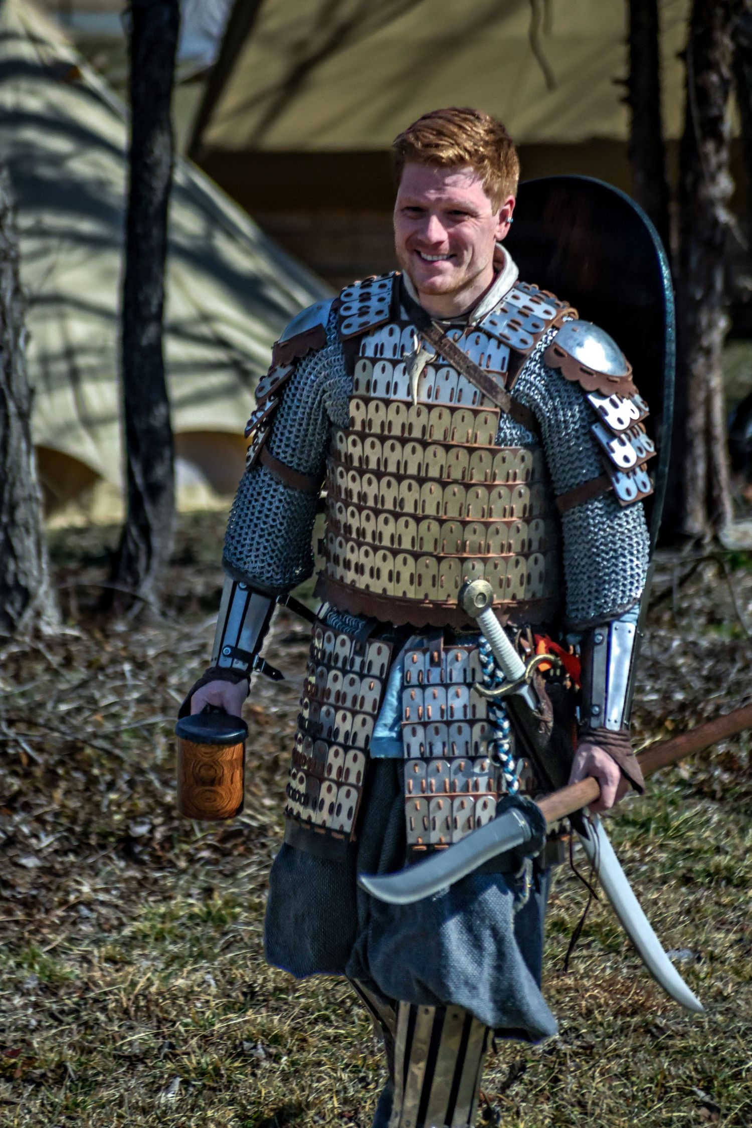 Medieval warrior wearing layered chainmail and scale armor holds a sword and drinking vessel in a forest setting.