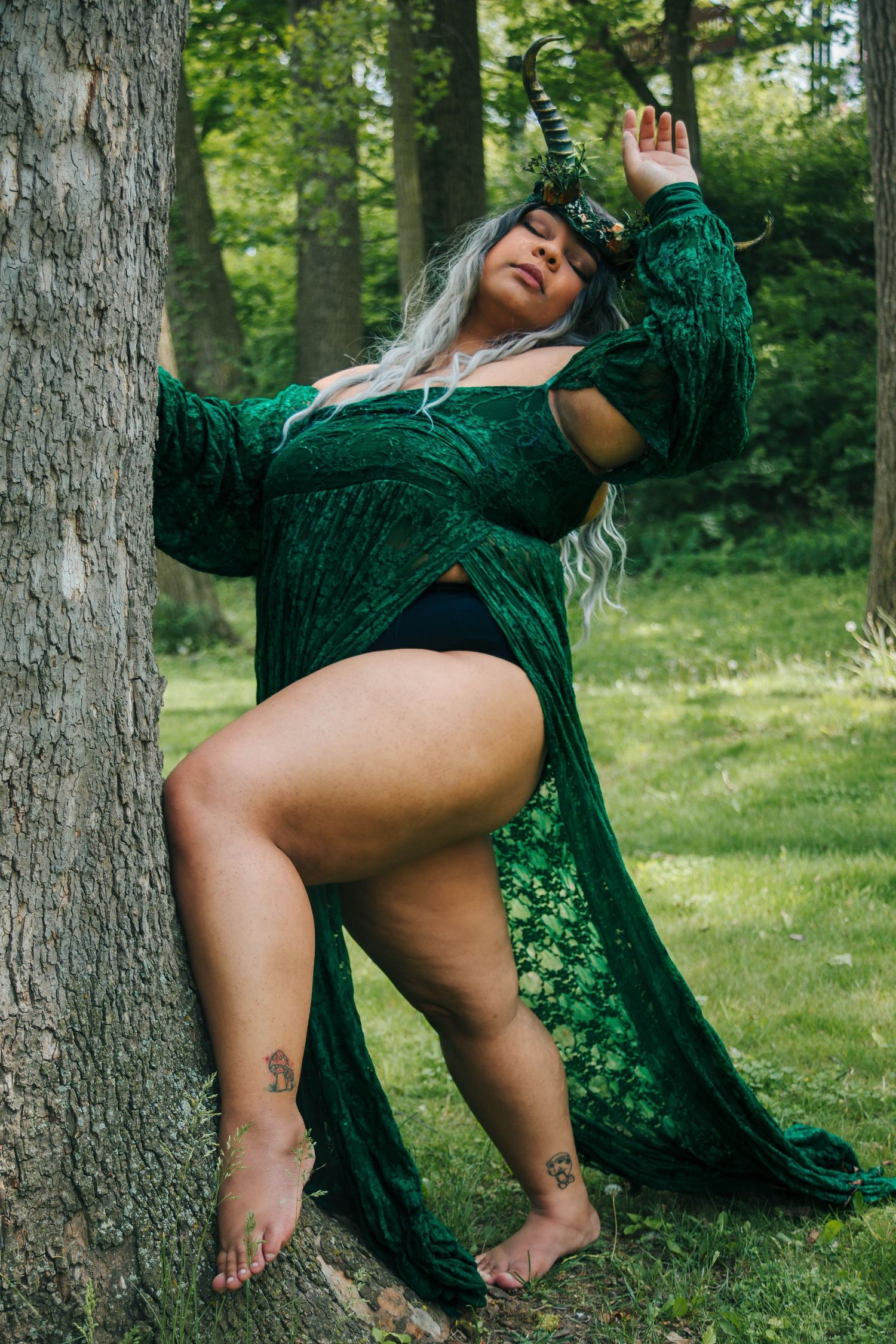 Woman in green velvet robe with fantasy horns poses against tree in forest setting.