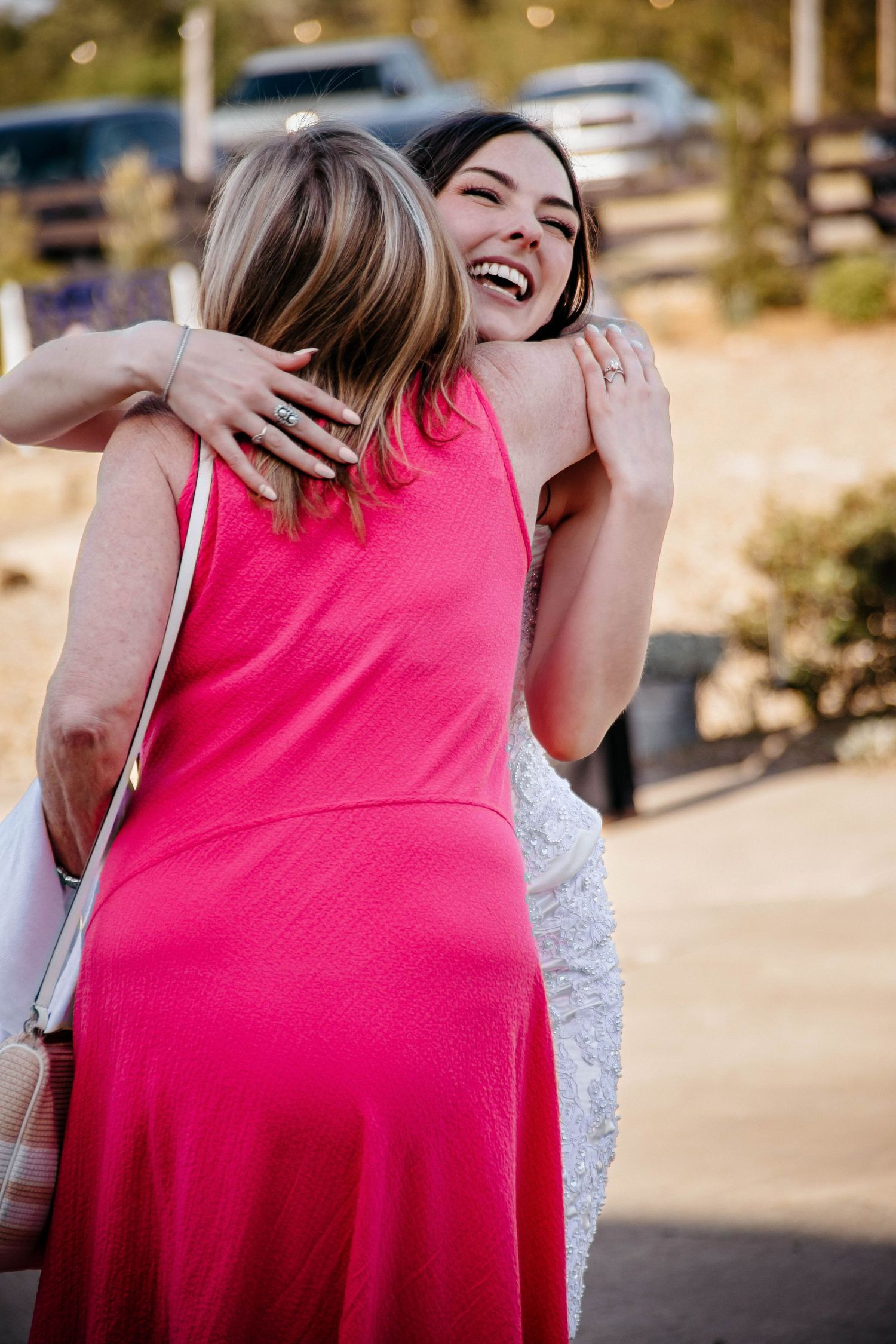 Two women in pink dresses share a joyful hug outdoors, one laughing warmly in a bright pink halter dress.
