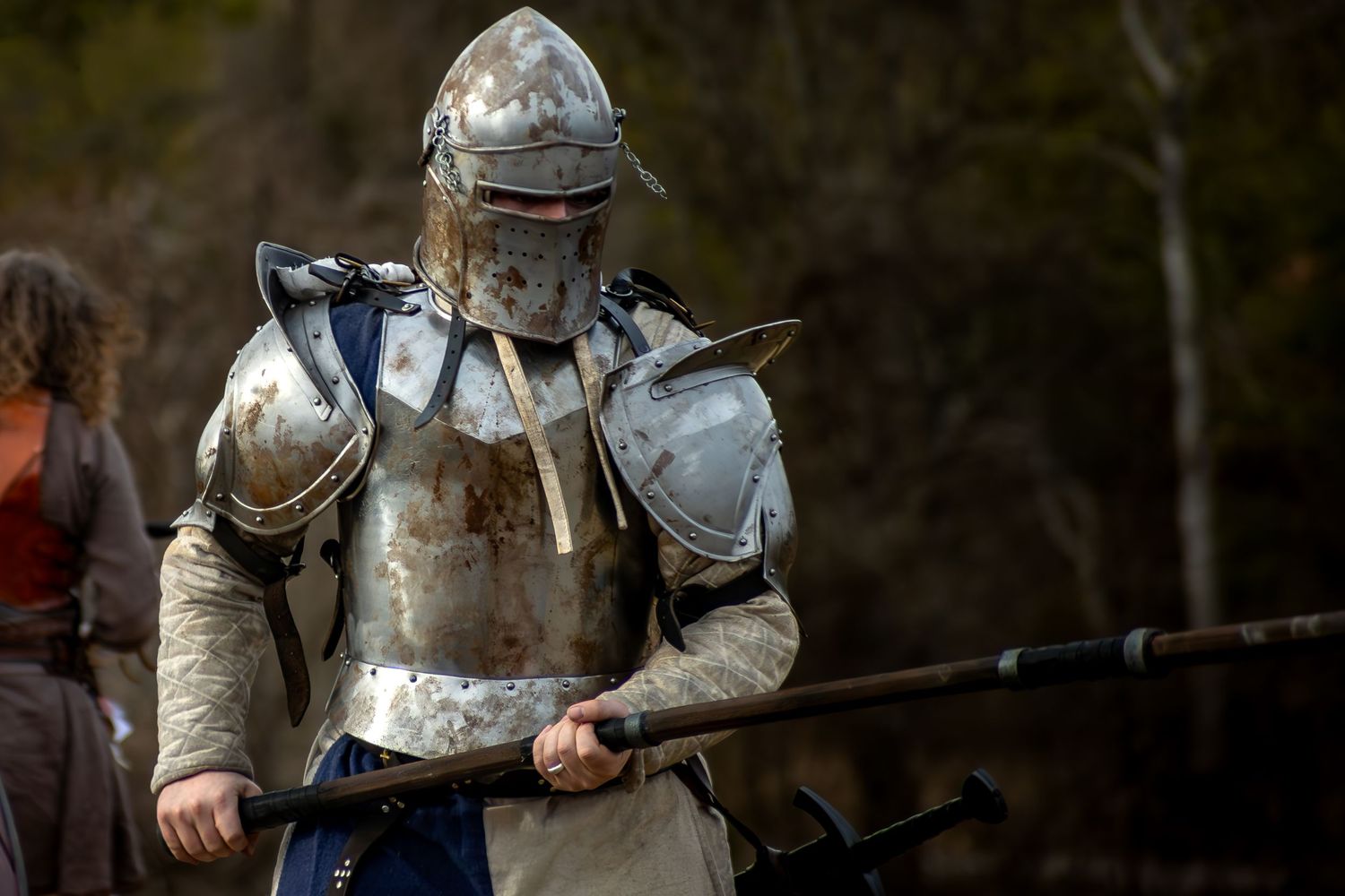 Medieval knight in battle-worn silver armor holds a spear during historical reenactment event.