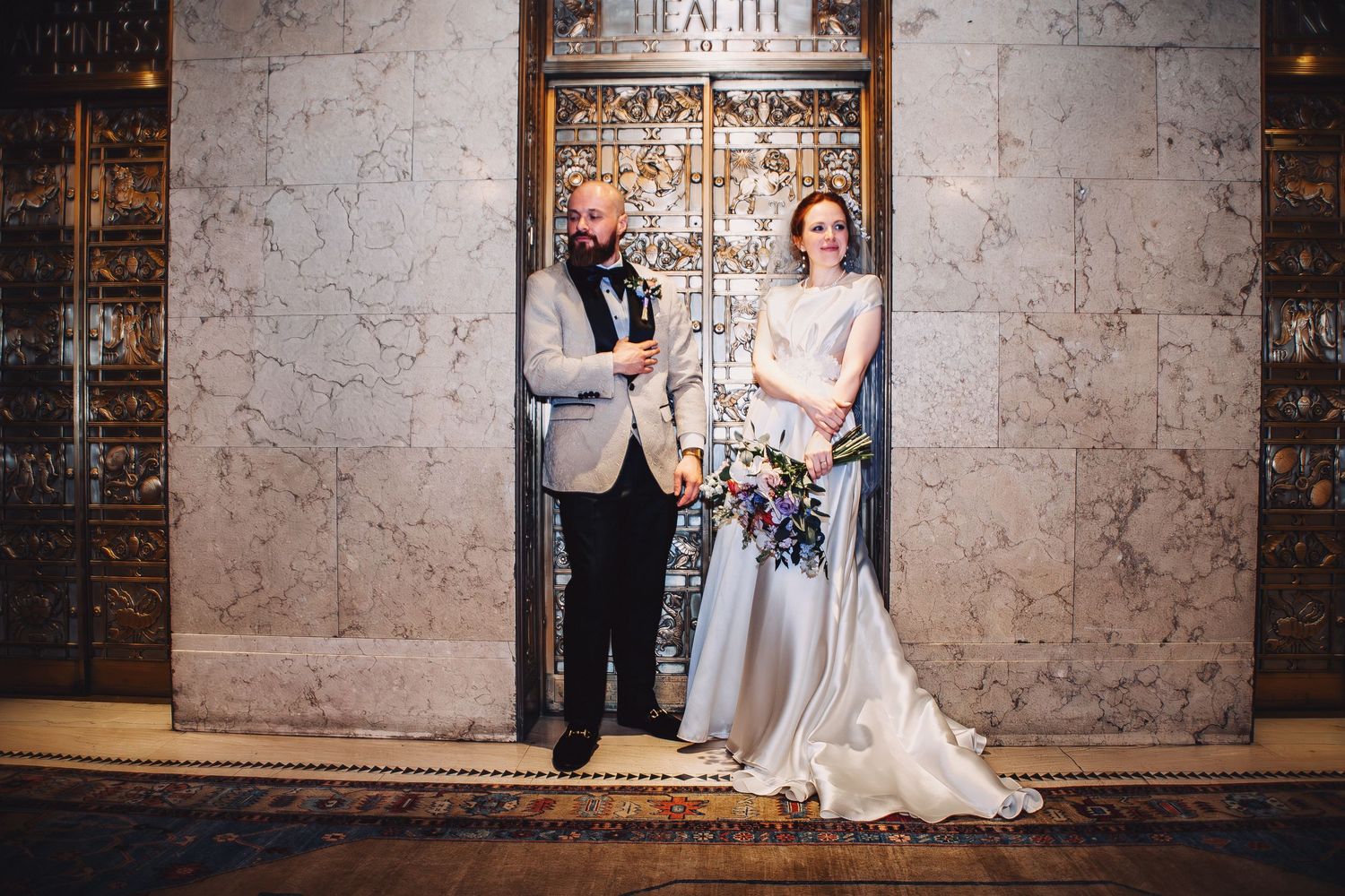 Bride and groom posing elegantly in front of ornate gold doors in a marble-walled venue on their wedding day.