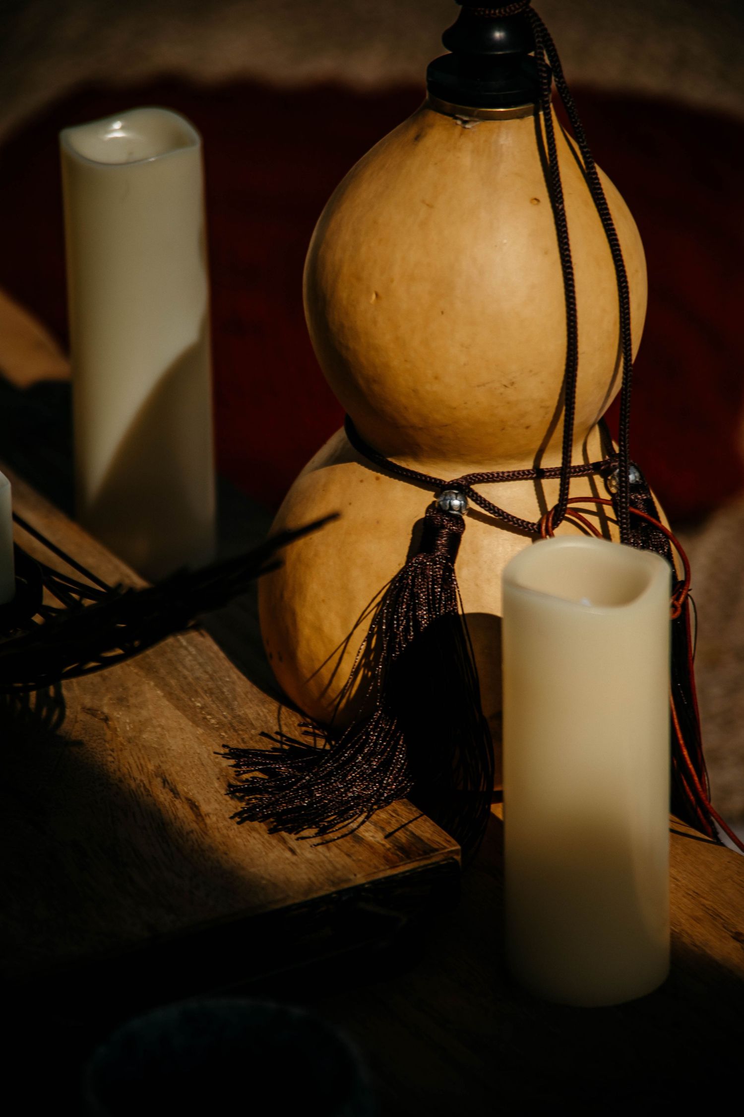 A decorative gourd with black tassels sits between two white pillar candles in a moody, atmospheric setting.